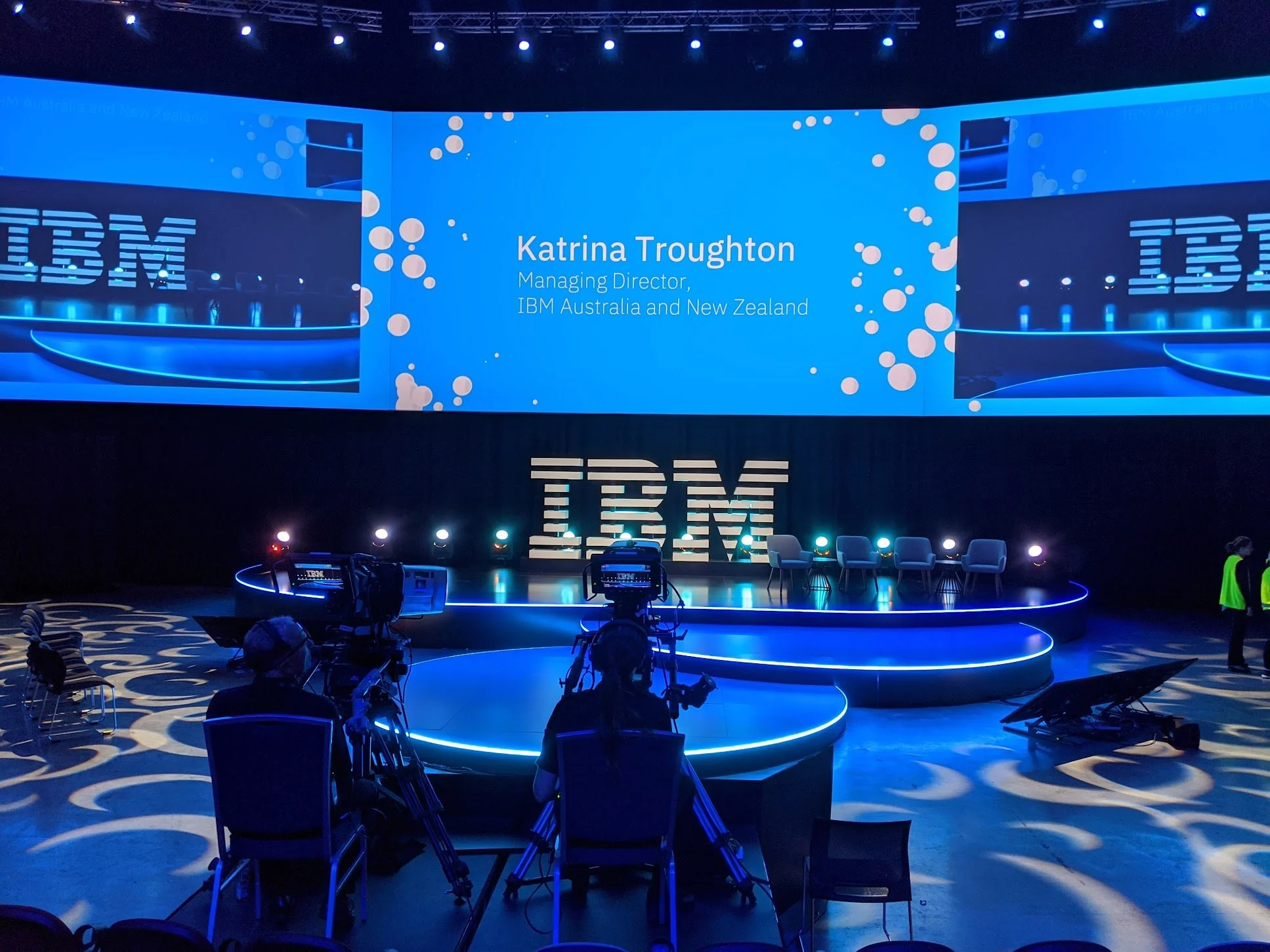 Stage setup for a presentation with a large screen displaying 'Katrina Troughton, Managing Director, IBM Australia and New Zealand,' and a decorative 'IBM' sign below. Cameras and chairs are arranged in front of the stage, with additional chairs on t