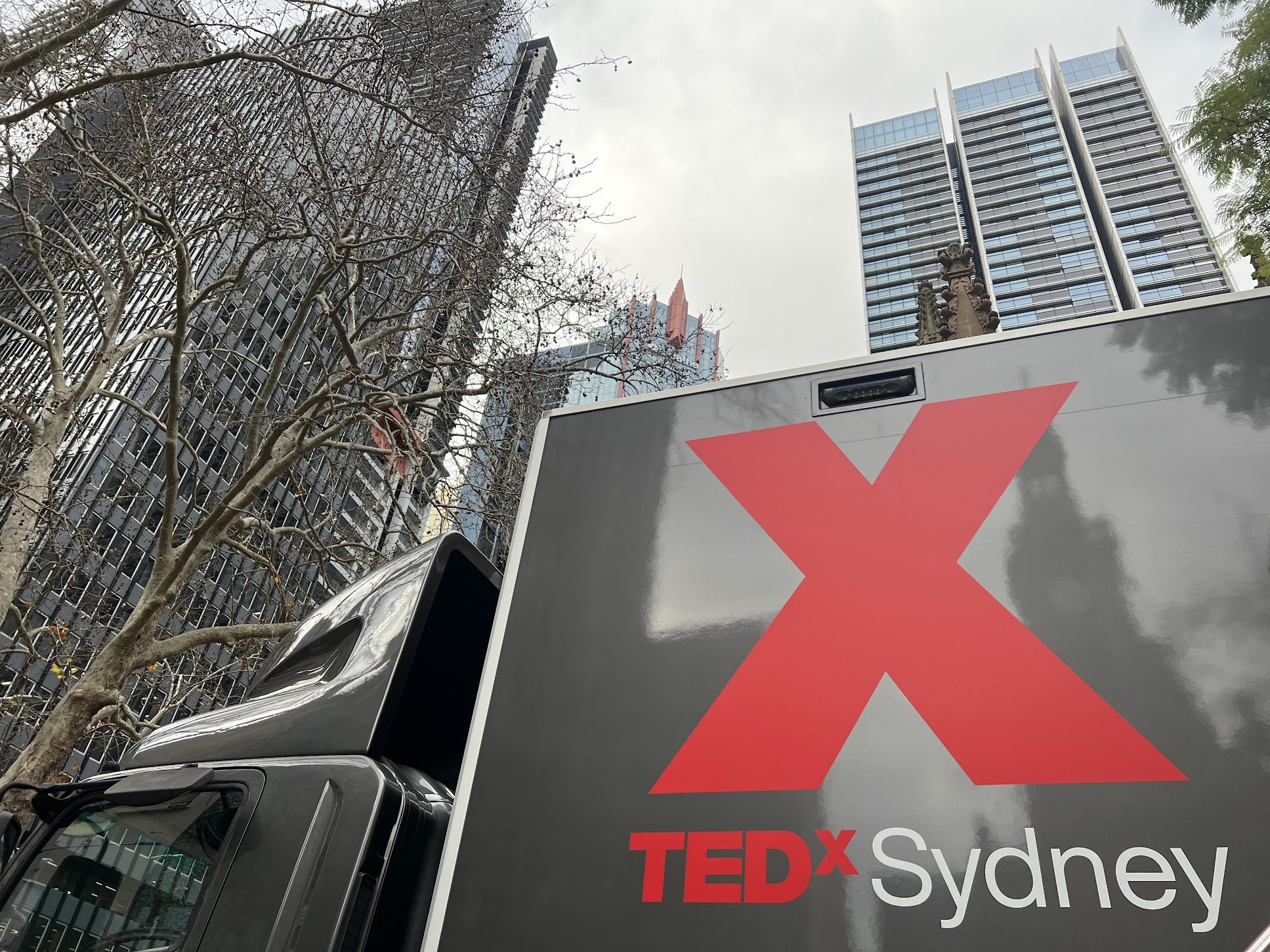 A truck with a large red X and TEDx Sydney logo on its side, set against a cityscape background with tall buildings and leafless trees.