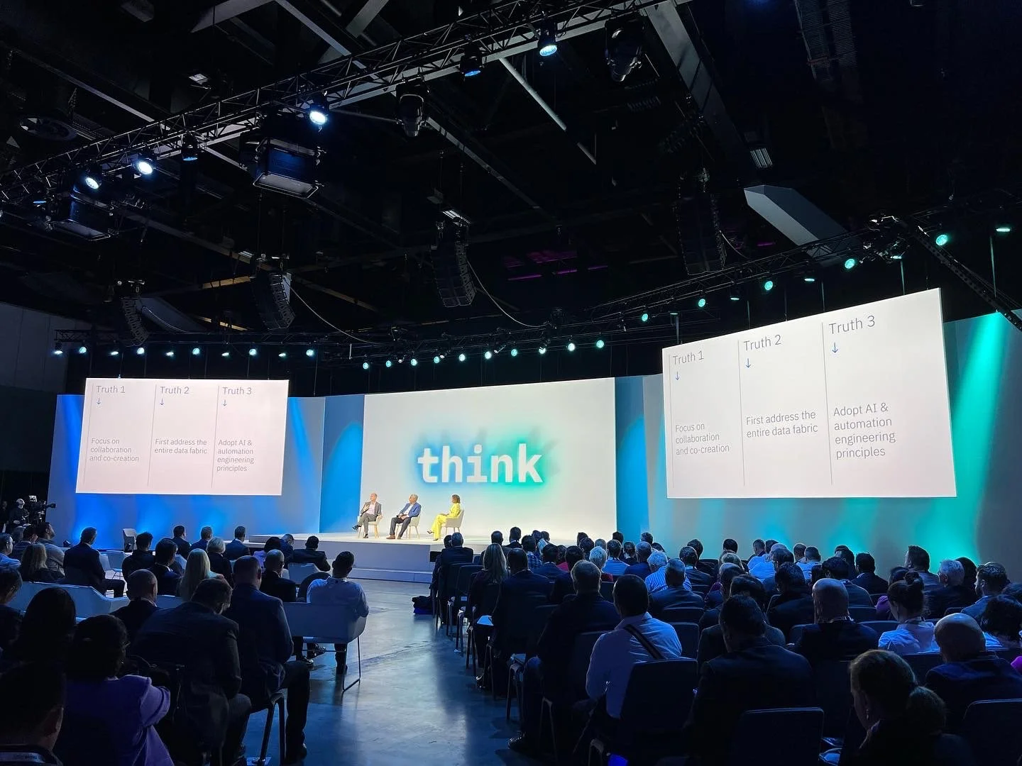 A large conference room with many attendees seated, facing a stage where a panel of three speakers is seated. The background displays the word 'think' in large letters, and screens on either side of the stage show presentation slides with three colum