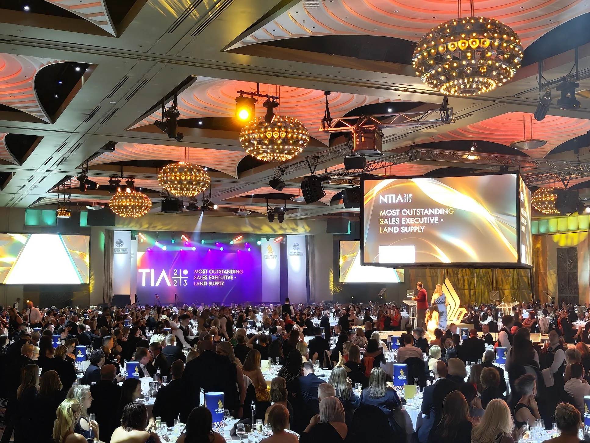 Large conference room filled with attendees at tables, stage with large screens displaying awards and presentation text, ornate ceiling lights.