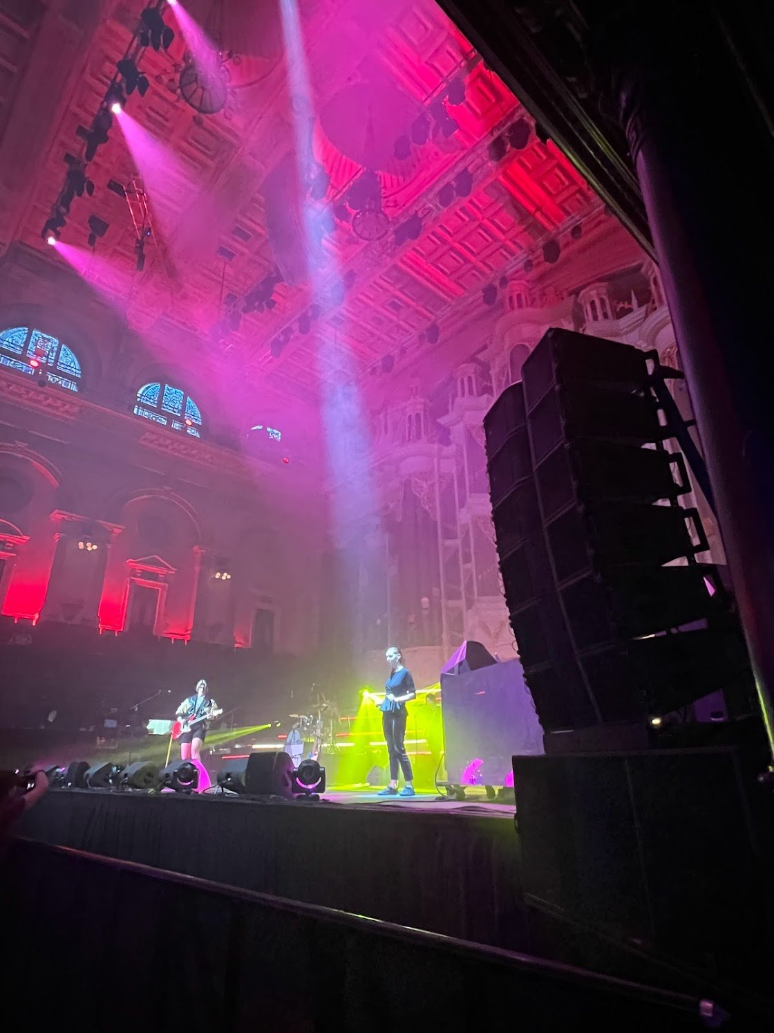 A live music performance on a stage inside a grand, ornate theater with pink and purple lighting, featuring musicians with guitars and microphones.