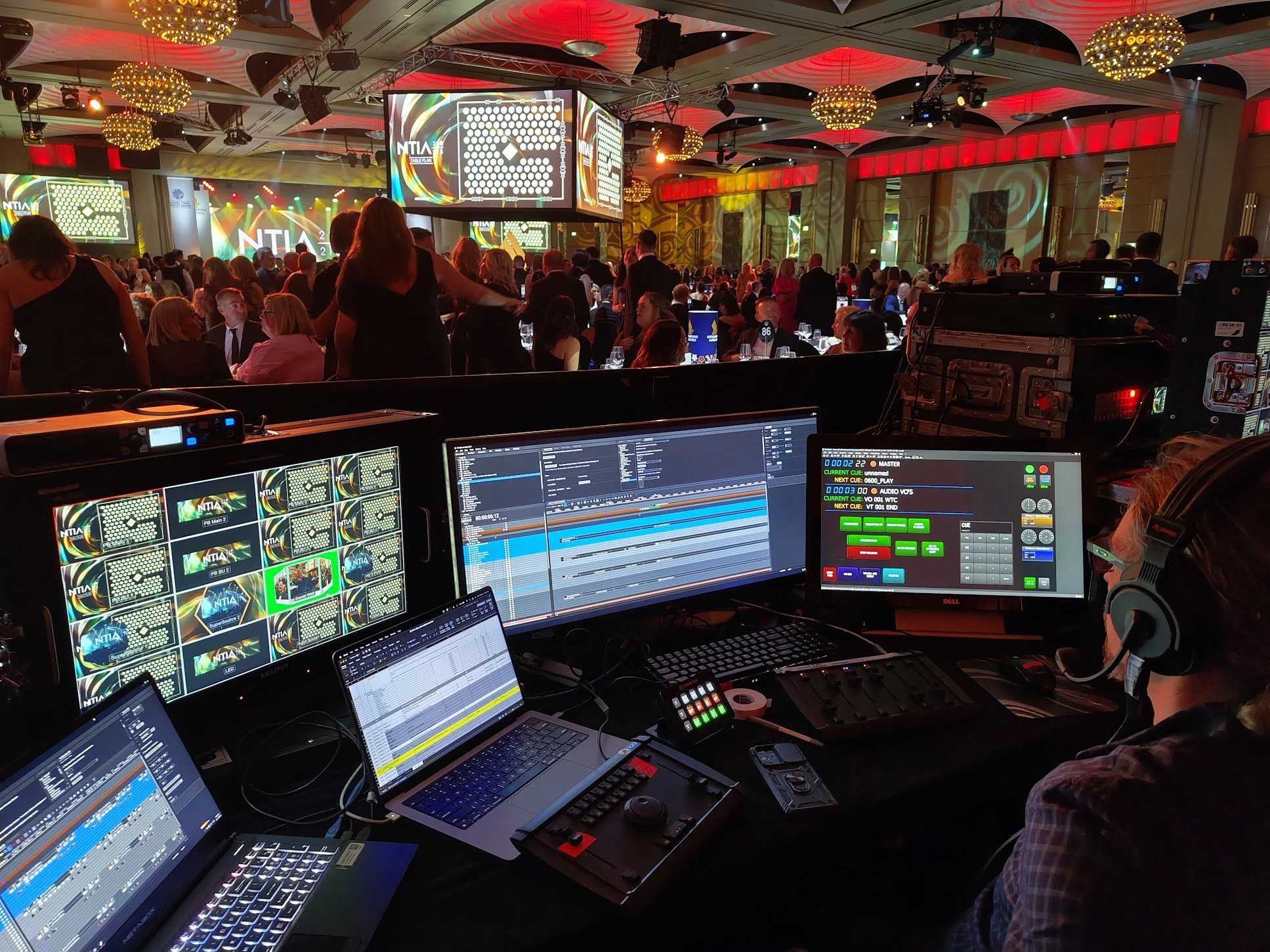 Photo of a large conference or award show with many attendees seated at round tables in a decorated ballroom. A large stage with multiple screens displays the logo 'NTIA' and colorful graphics, and is brightly lit with red, yellow, and green lights. 