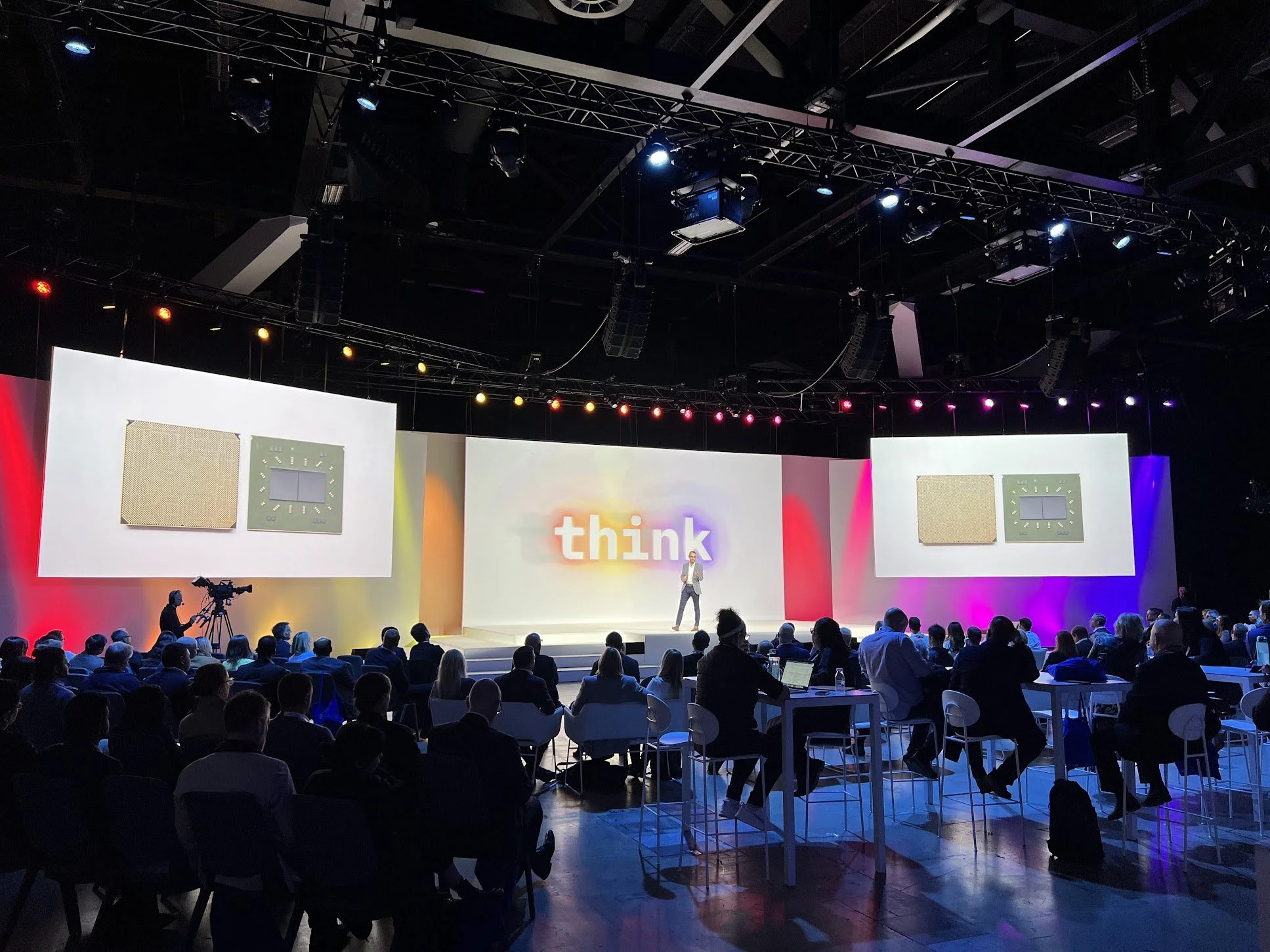A conference or presentation event in a large auditorium with a stage and big screens, with a speaker presenting to an audience seated at tables and in chairs; the stage has colorful lighting and the word 'think' displayed on the central screen.