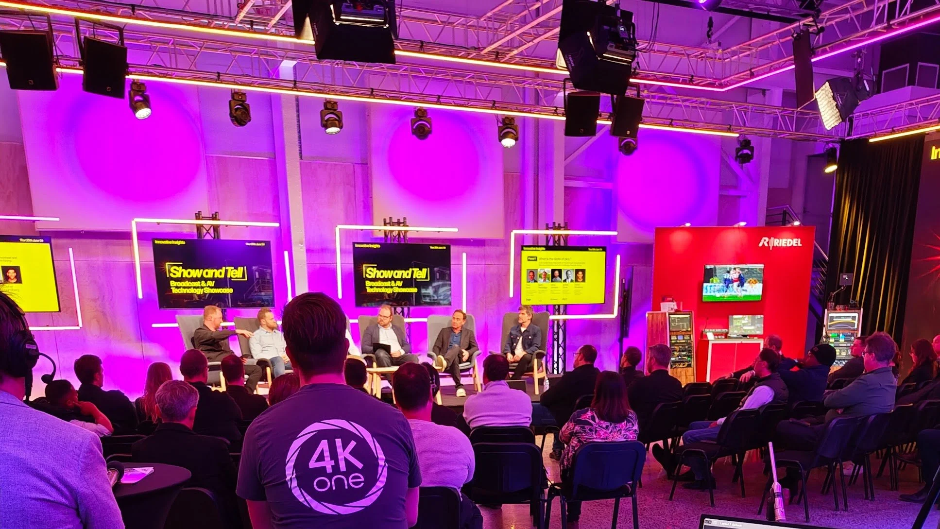 People attending a broadcast and TV technology showcase event, seated and watching a panel discussion on a stage with four individuals. The stage is lit with purple lighting, with screens and equipment visible behind the speakers.