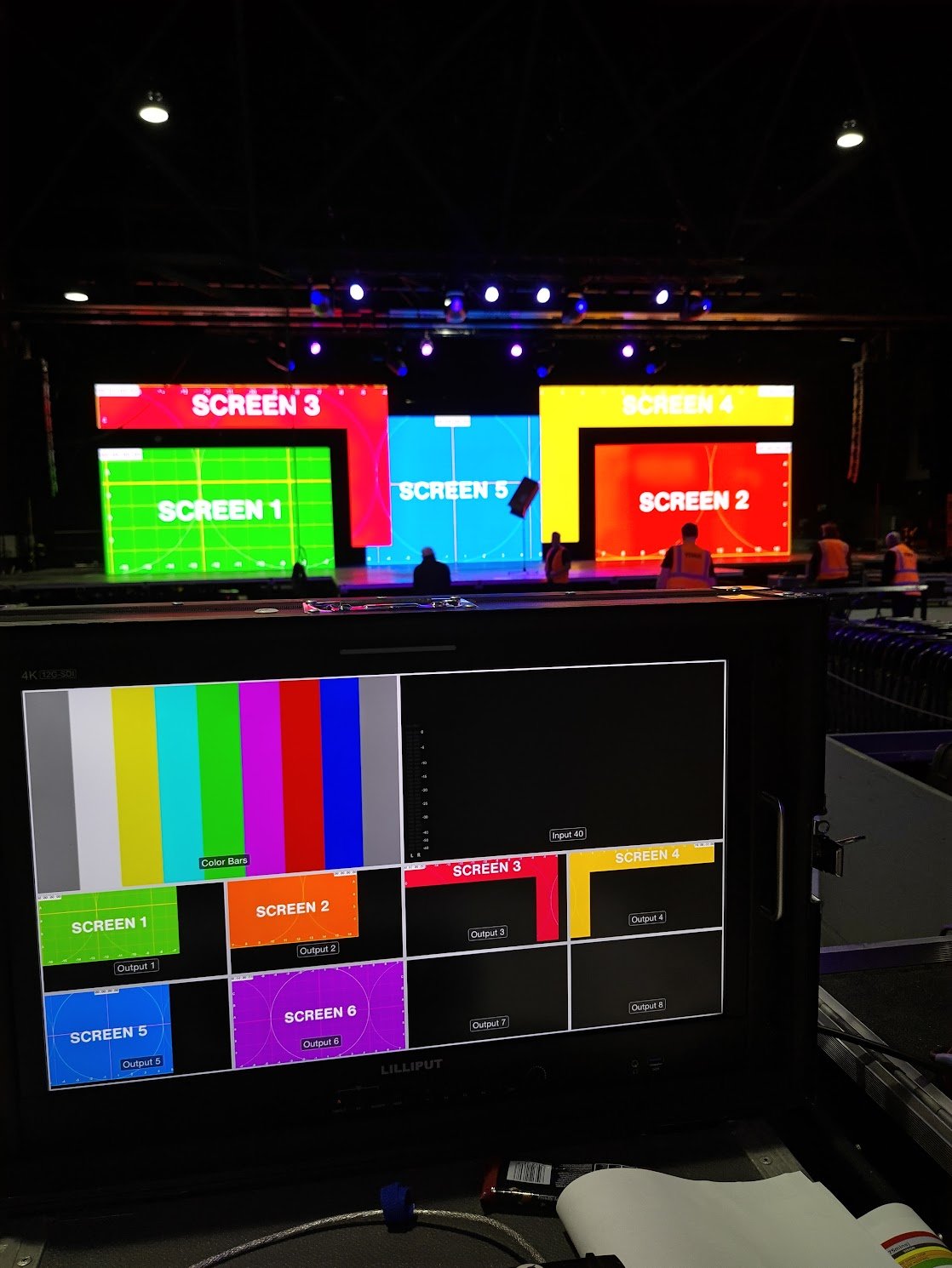 A stage with multiple screens, labeled Screen 1 through Screen 5, in a dark auditorium, with a control monitor showing color test bars and layouts for the screens.