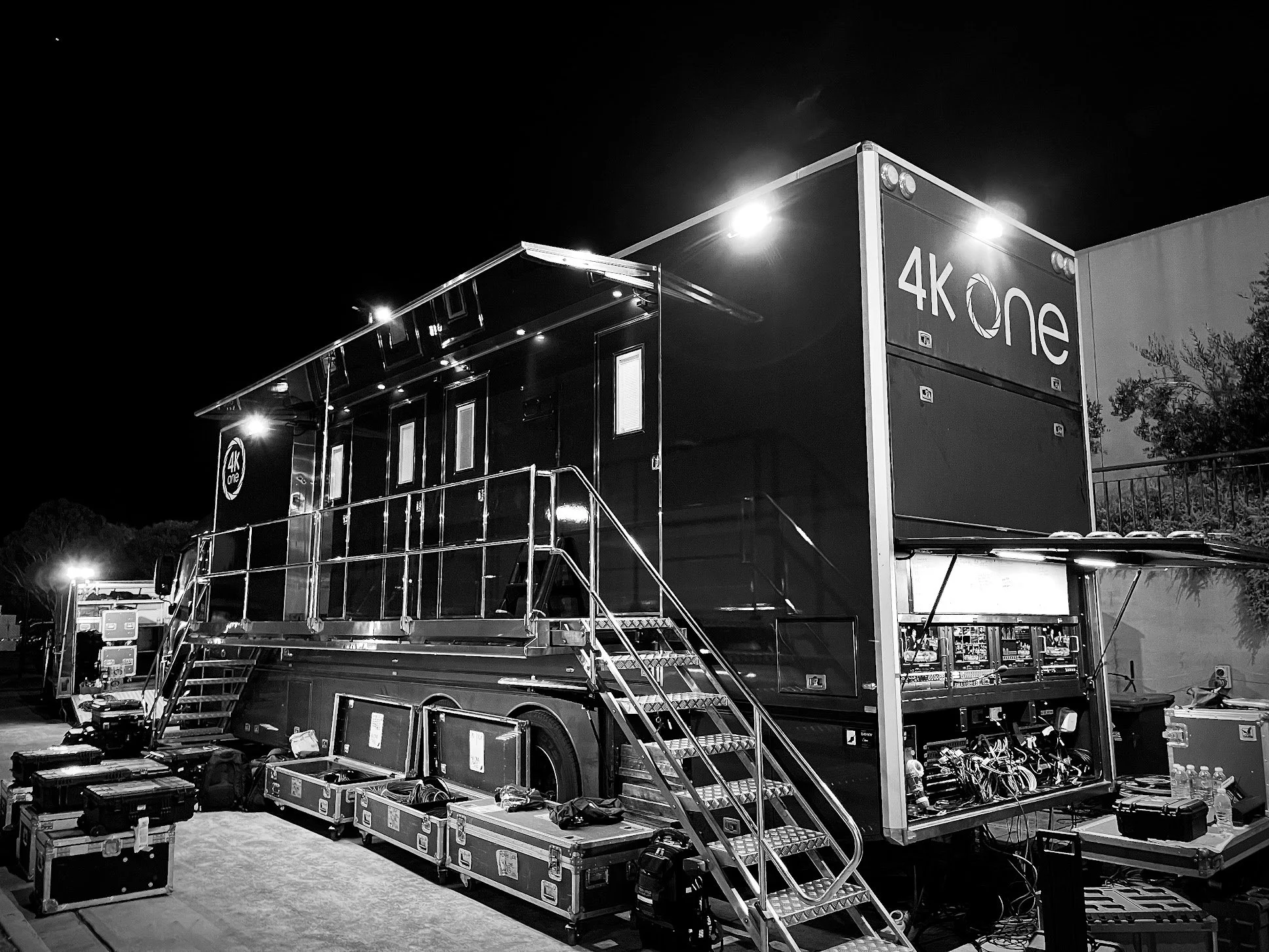 Black and white photo of a large mobile production truck at night, labeled '4K one,' with equipment cases around it and stairs leading to the entry.
