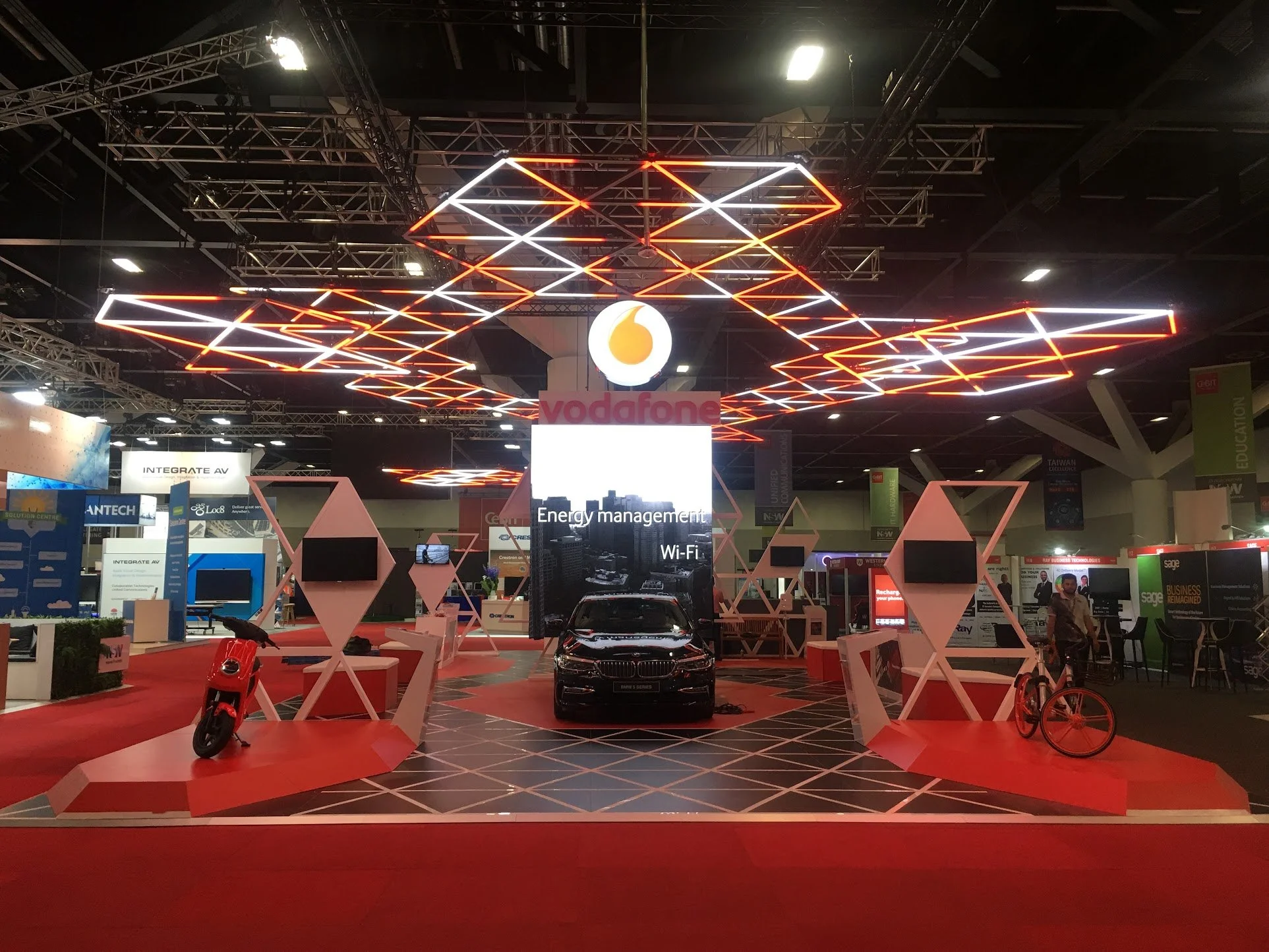 Exhibition booth featuring a black car, red and pink geometric structures, a scooter, and a bicycle on a red carpet with bright neon lighting overhead, branded by Vodafone.