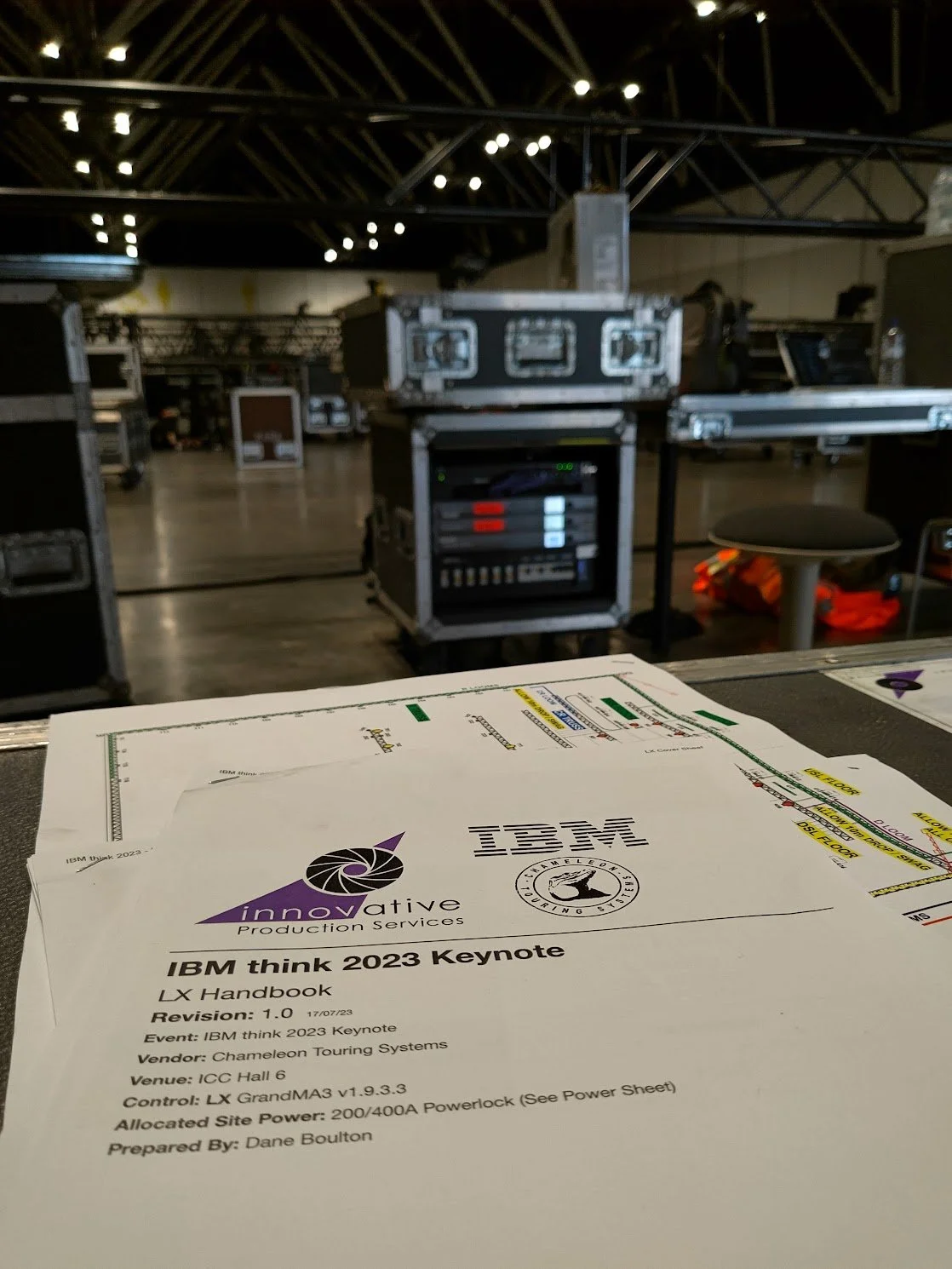 Document titled 'IBM think 2023 Keynote' on a table with event details, with a background of equipment, racks, and a table with a water bottle, in a large conference or event hall.