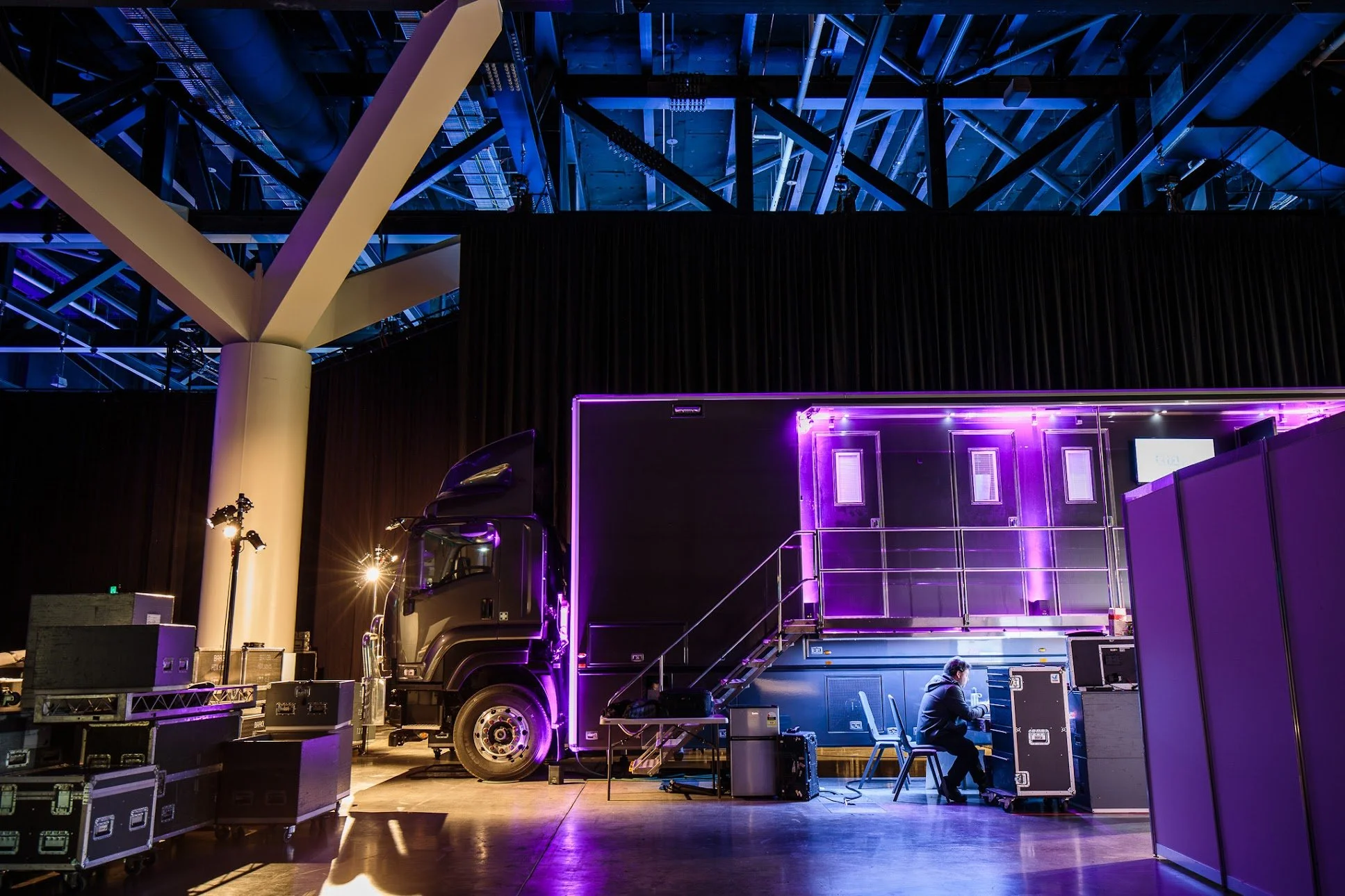 A large black truck is parked inside a venue with a high ceiling and exposed metal beams illuminated in blue. To the right of the truck, a person is working at a desk with electronic equipment, surrounded by purple lighting and black storage cases.