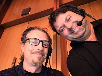 Two men wearing headsets and glasses, smiling for a selfie inside a wooden-paneled room.