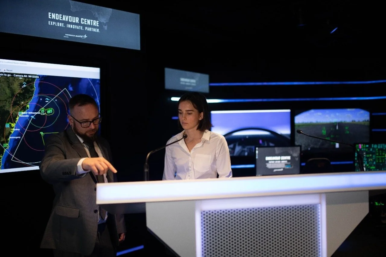 Two people, a man and a woman, stand at a podium in a dark room with multiple computer screens and monitors displaying maps and flight simulation graphics behind them. The room is labeled 'Endeavour Centre'.
