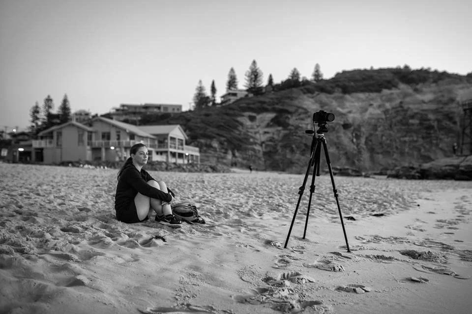A woman sitting on the sandy beach near a tripod with a camera, overlooking the ocean with houses in the background