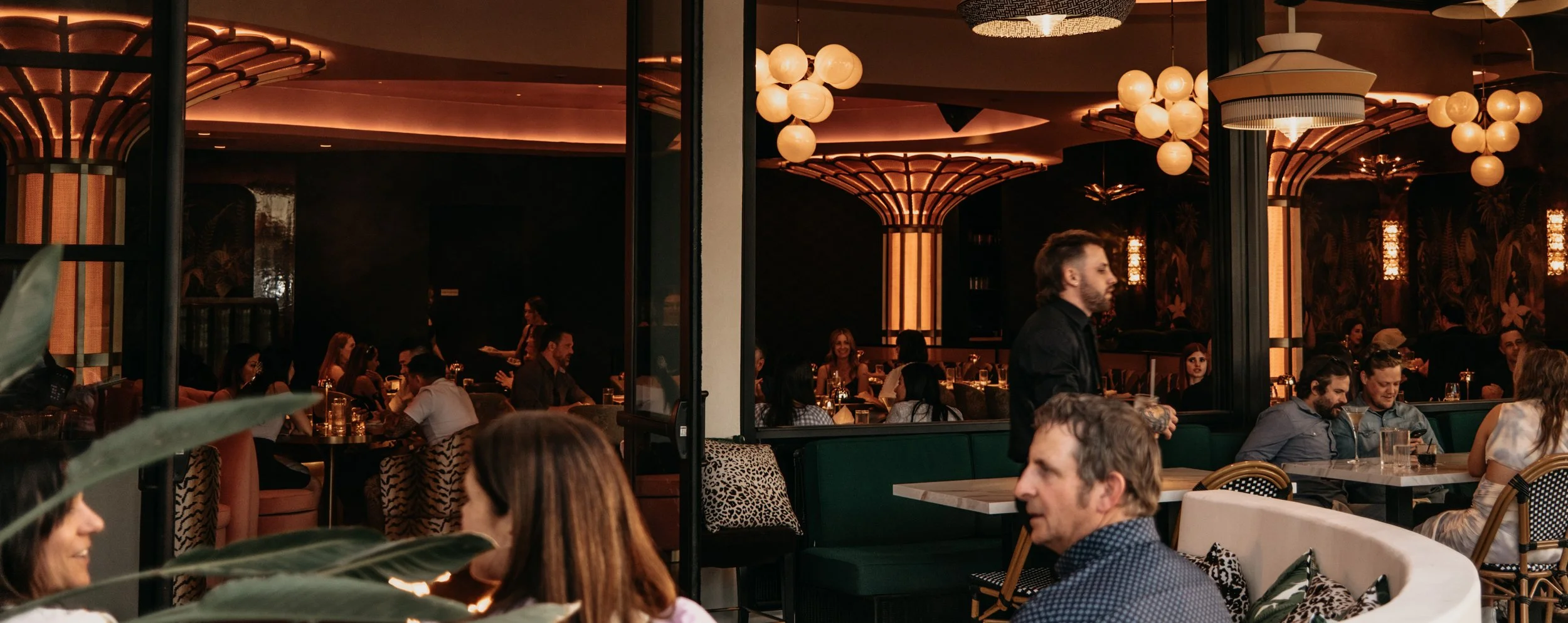 Interior of a dimly lit upscale restaurant with decorative lighting, multiple diners seated at tables, and a waiter serving. The decor features warm tones, modern chandeliers, and lush tropical wall coverings.
