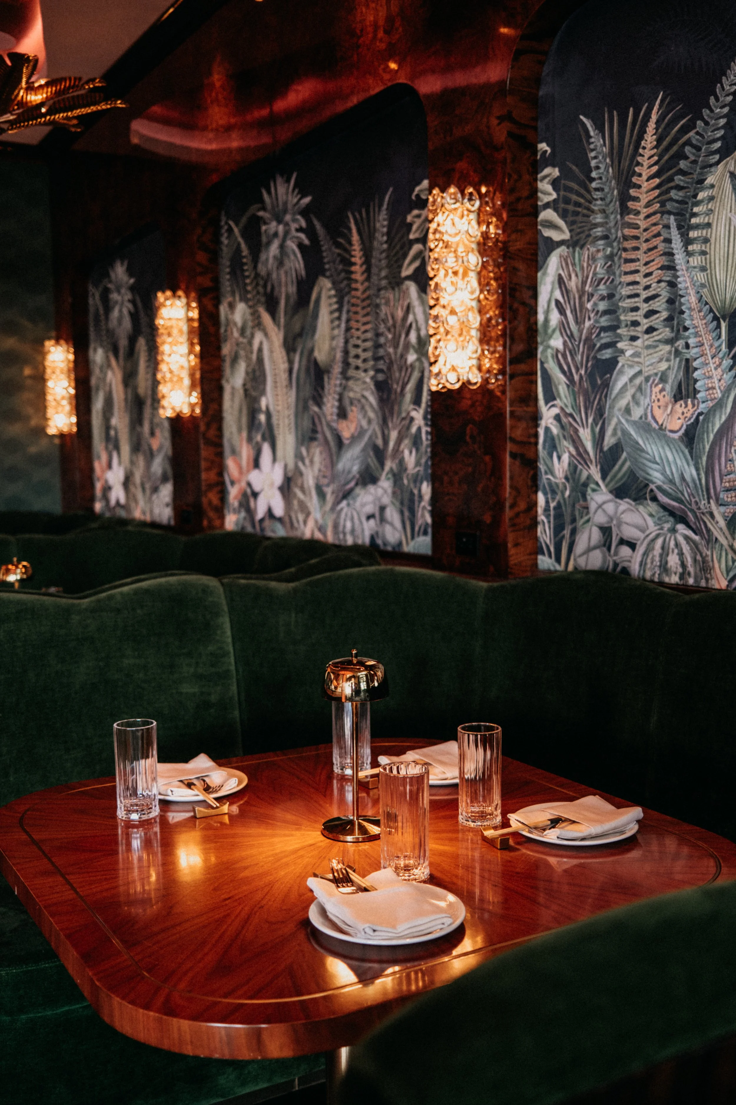 A restaurant table set for four with napkins, forks, glasses, and a small lamp, surrounded by green velvet booths and decorated with tropical patterned wallpaper and wall-mounted lights.