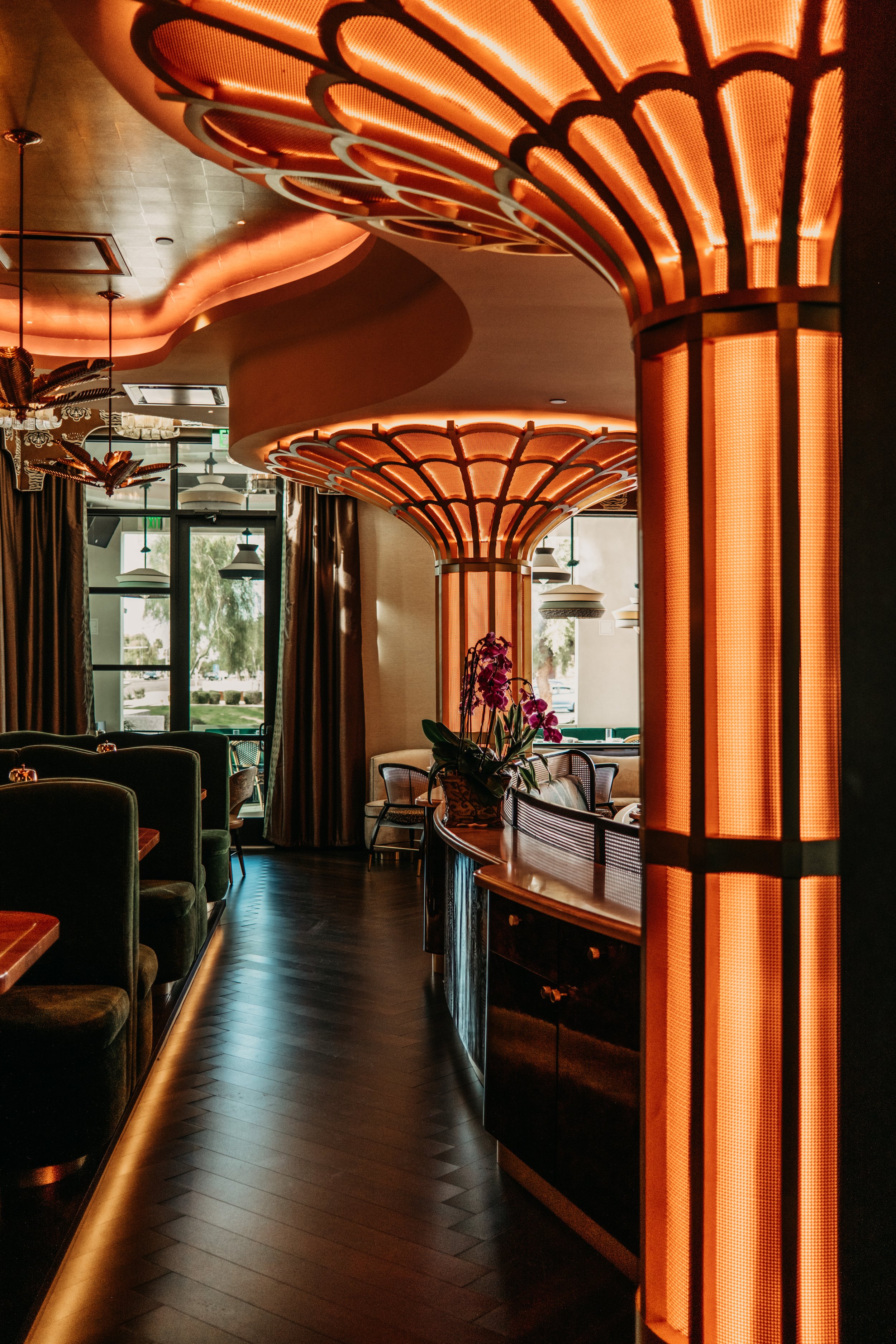 Interior of a restaurant with illuminated Art Deco style orange lighting fixtures, dark wood flooring, green velvet booths, and large windows with outdoor views.