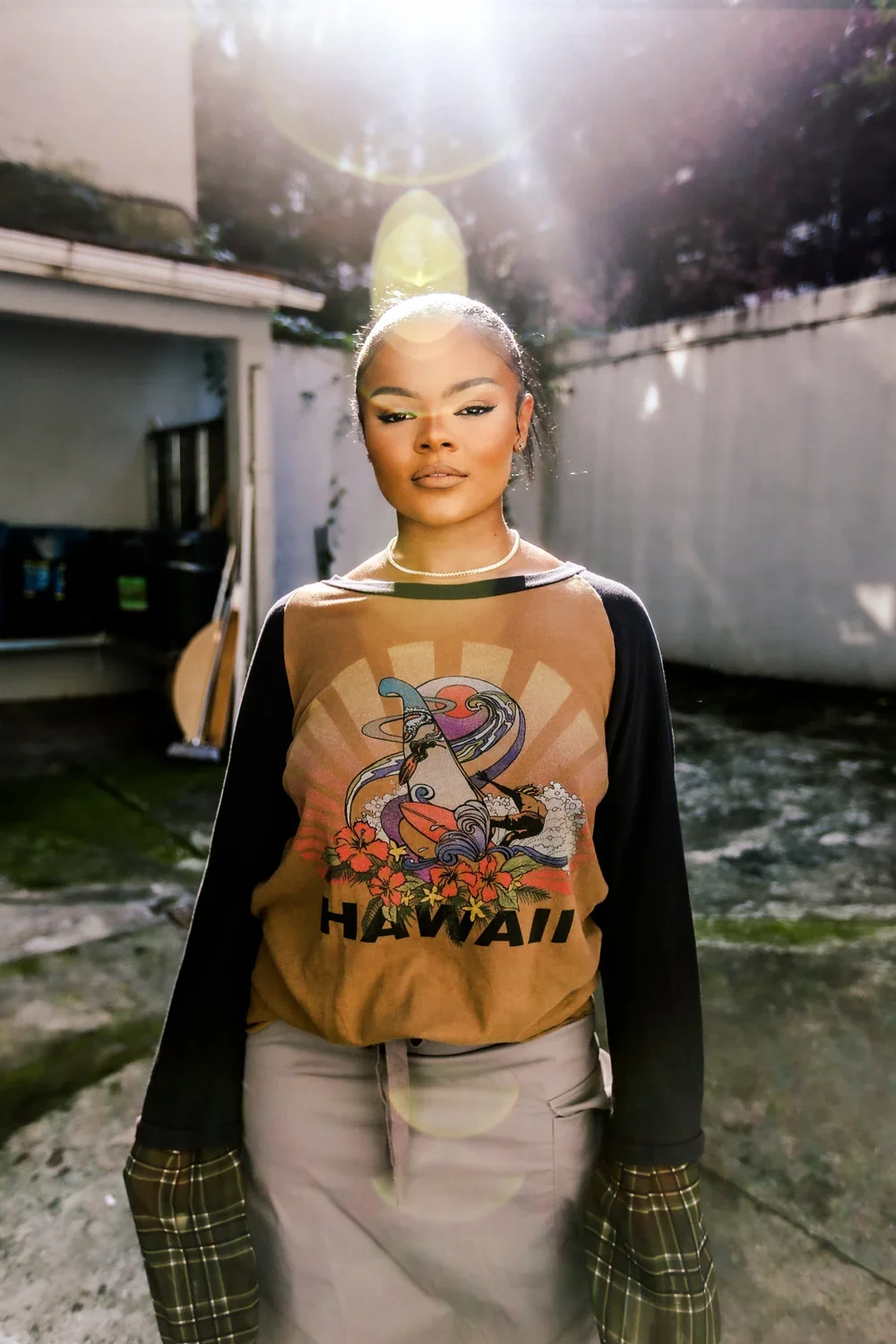 A young woman standing outdoors in sunlight wearing a Hawaii-themed sweatshirt with a graphic of a wave, a surfboard, and flowers, paired with light pants and plaid sleeves.