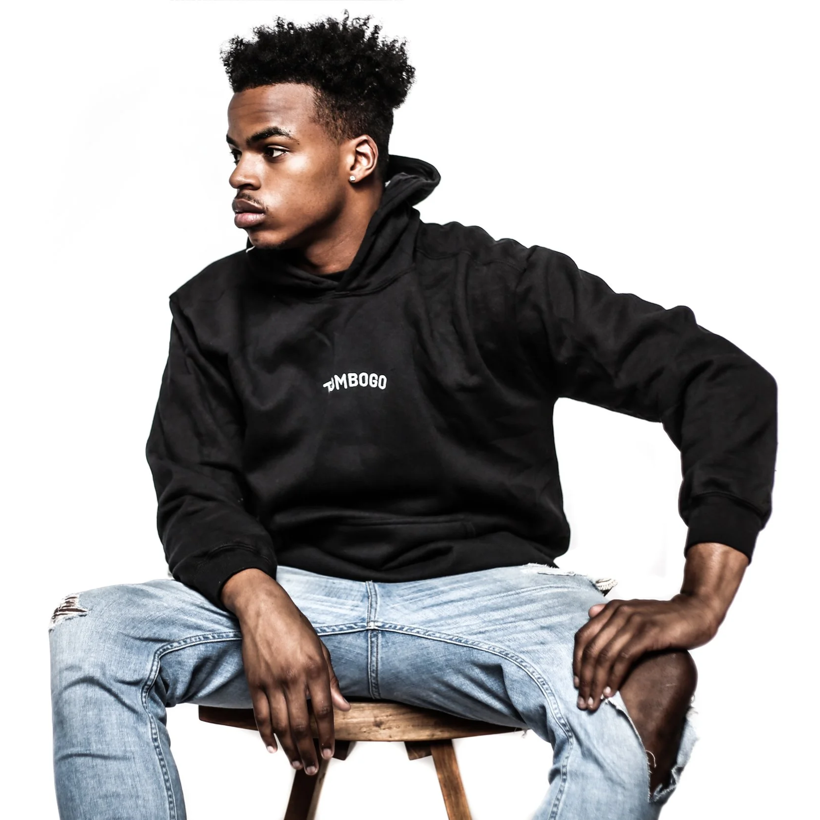 Young man with dark skin and curly hair sitting on a wooden stool, wearing a black hoodie with the word TMBOGO on it and ripped light blue jeans, against a plain white background.