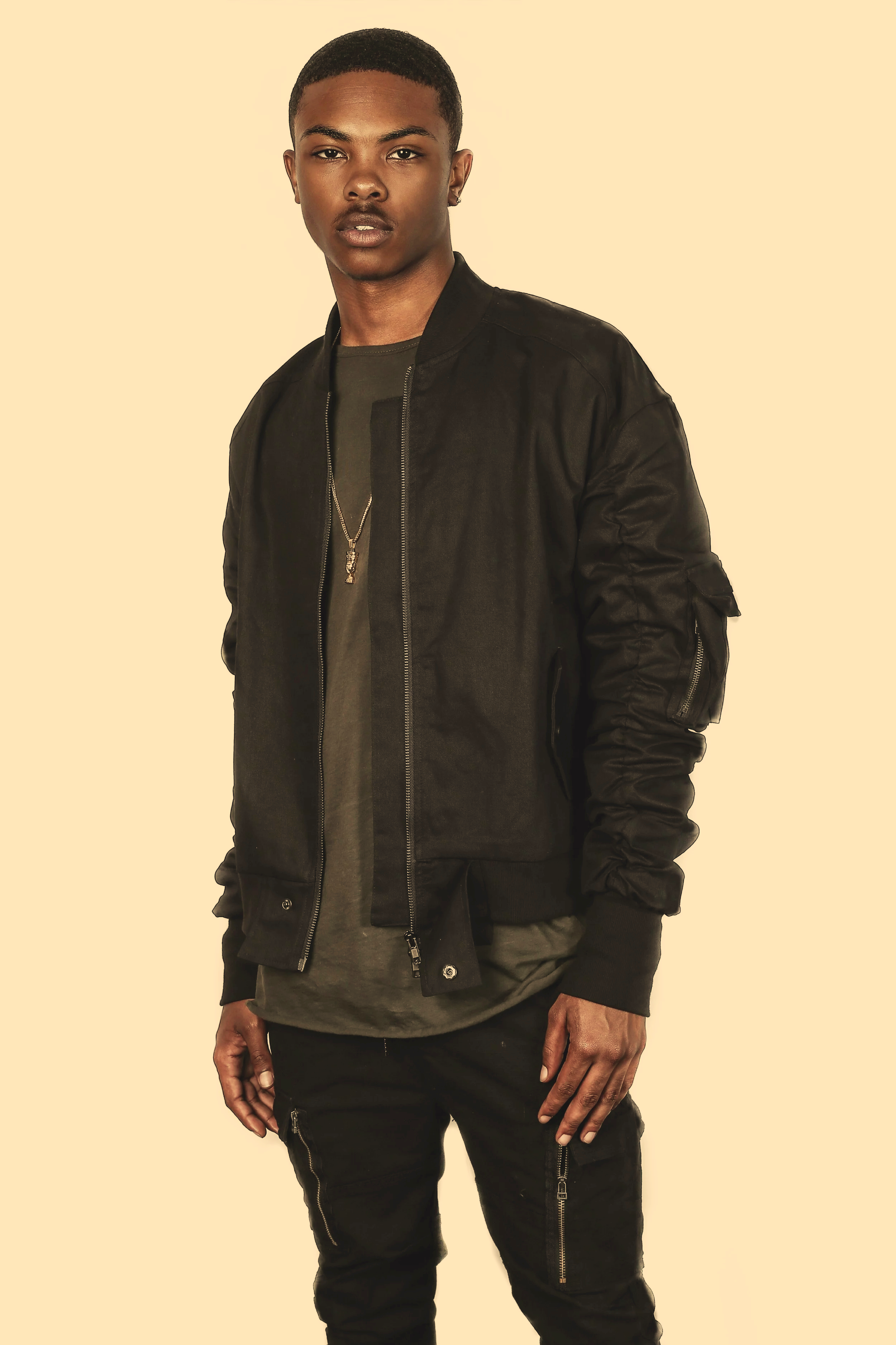 A young man with dark skin and short hair standing against a plain light-colored background, wearing a black bomber jacket, dark pants with zippers, and a necklace.