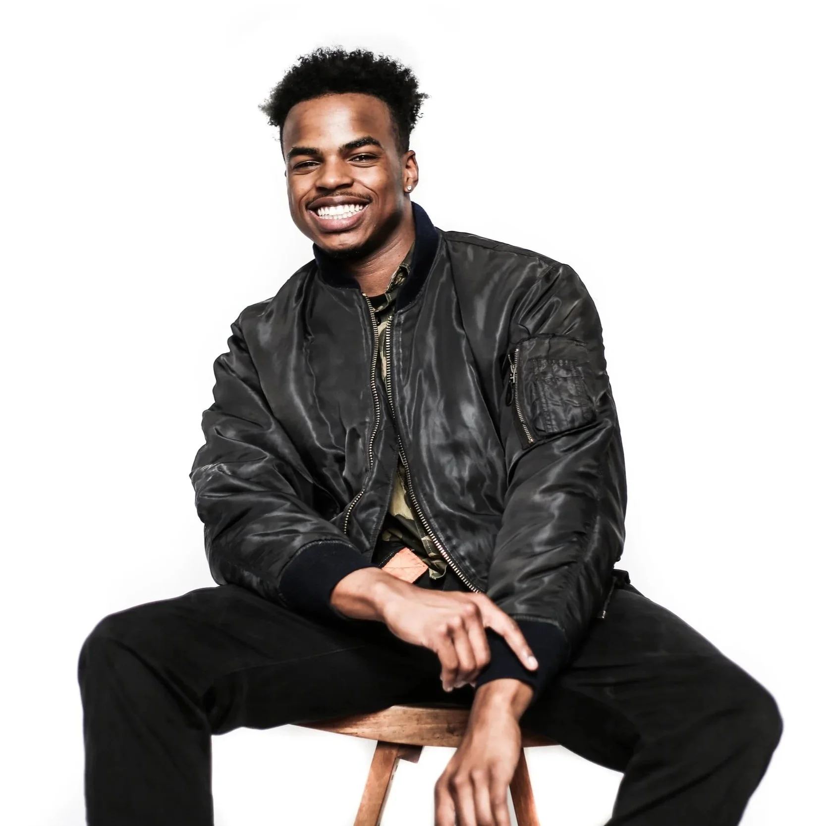 Young man smiling, sitting on a stool against a white background, wearing a black bomber jacket and black pants.