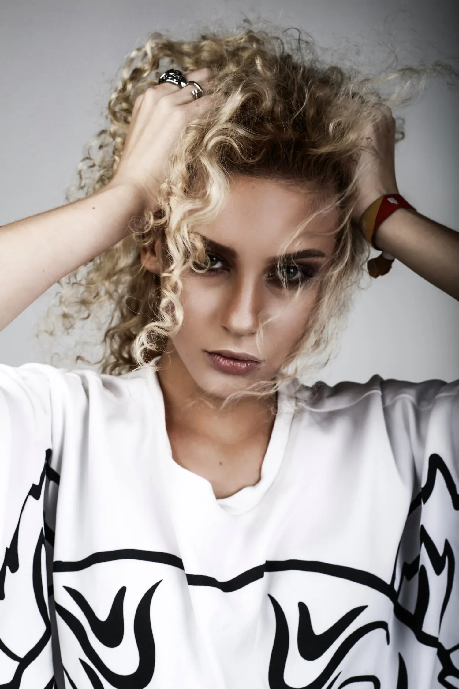 Young woman with curly blonde hair, wearing a white shirt with black graphic design, looking into the camera with hands in her hair.