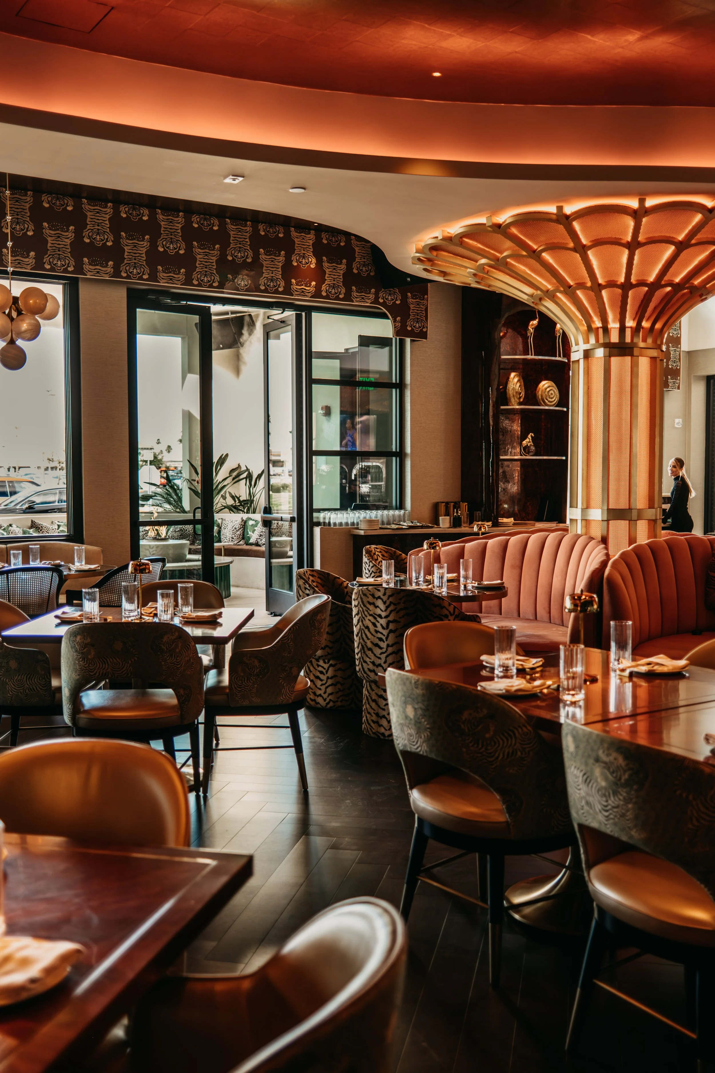 Interior of a stylish restaurant with warm lighting, patterned chairs, round tables set with glasses and napkins, and an open door leading to an outdoor patio with plants.