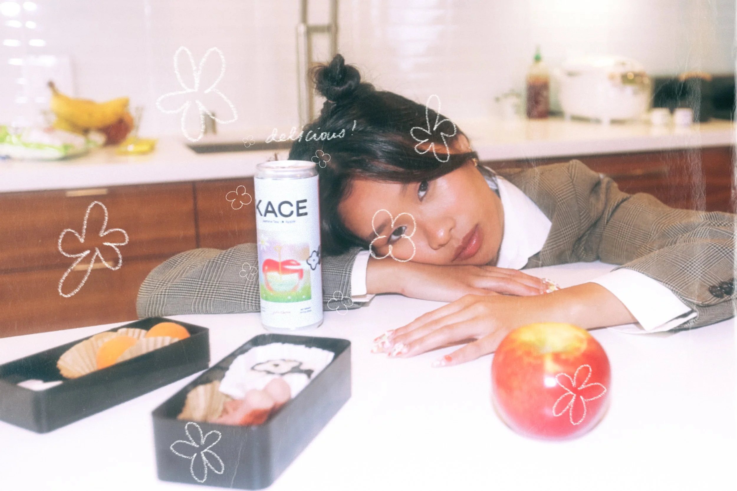 Woman with dark hair styled in a small bun, resting her head on her arms on a kitchen table with an apple and lunch boxes in front. There are decorative doodles of flowers and the word 'delicious' on the image.
