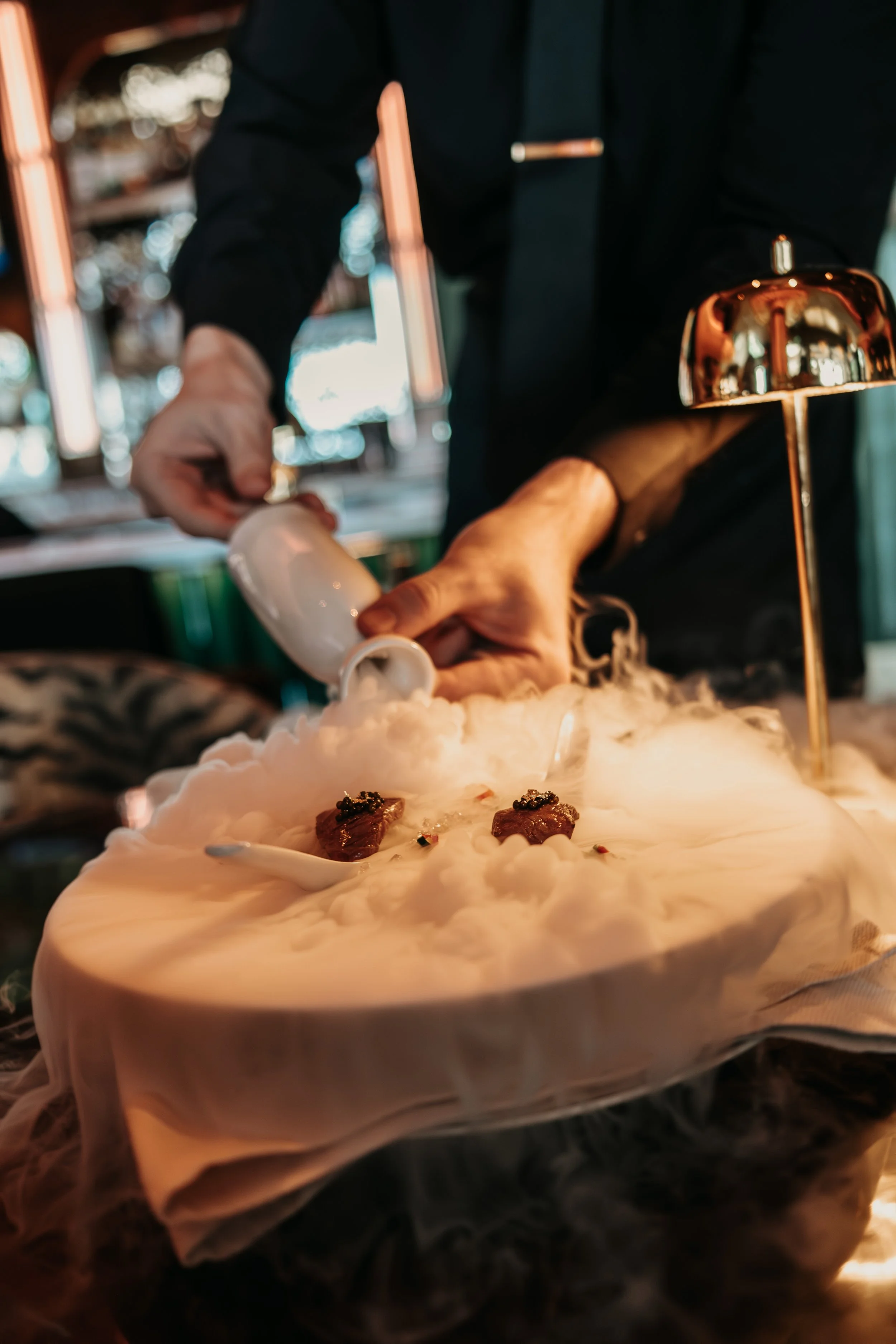 Chef pouring liquid nitrogen onto a dish with small pieces of meat, creating smoke and fog for a culinary presentation in a restaurant setting.