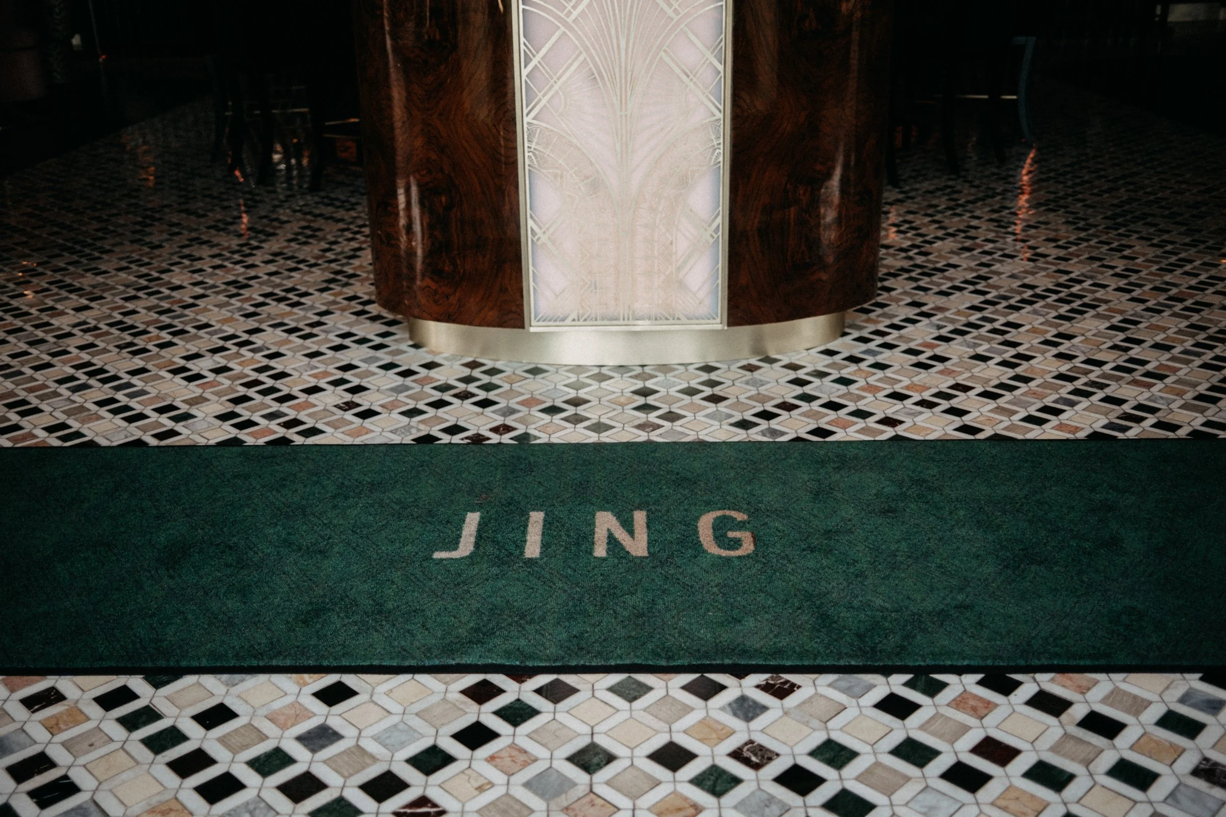 A green floor mat with the word 'JING' written on it in white letters, placed on a multicolored tile floor in front of a wooden counter with decorative glass panel.