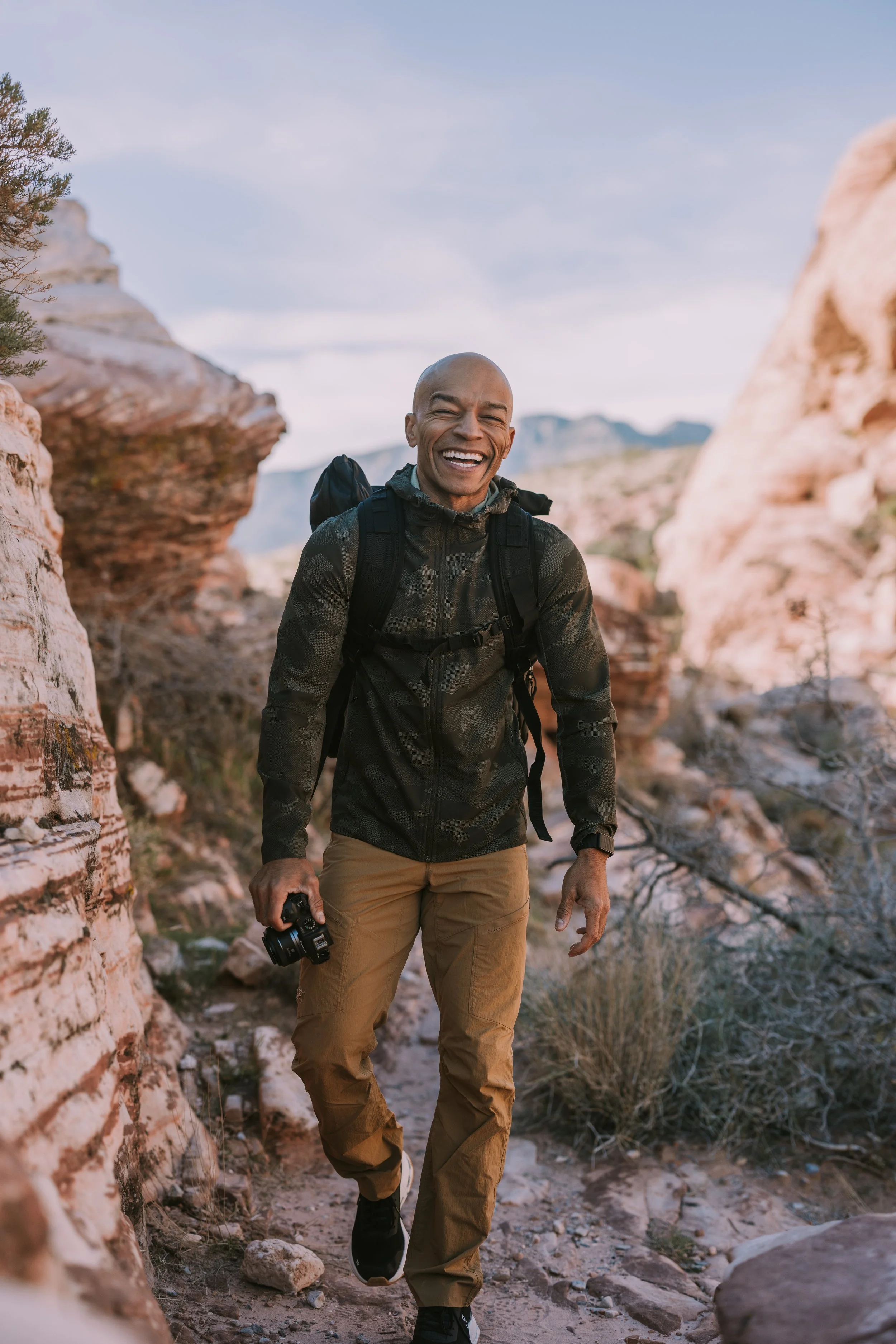 Man hiking in a desert canyon with rocks and sparse vegetation, smiling and holding a camera.