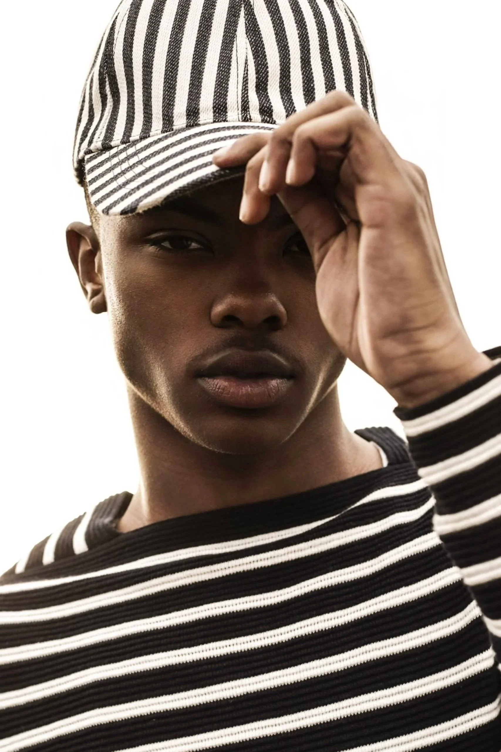 A young person with dark skin wearing a black and white striped baseball cap and a matching striped shirt, holding the brim of the cap with one hand, looking directly at the camera against a plain white background.