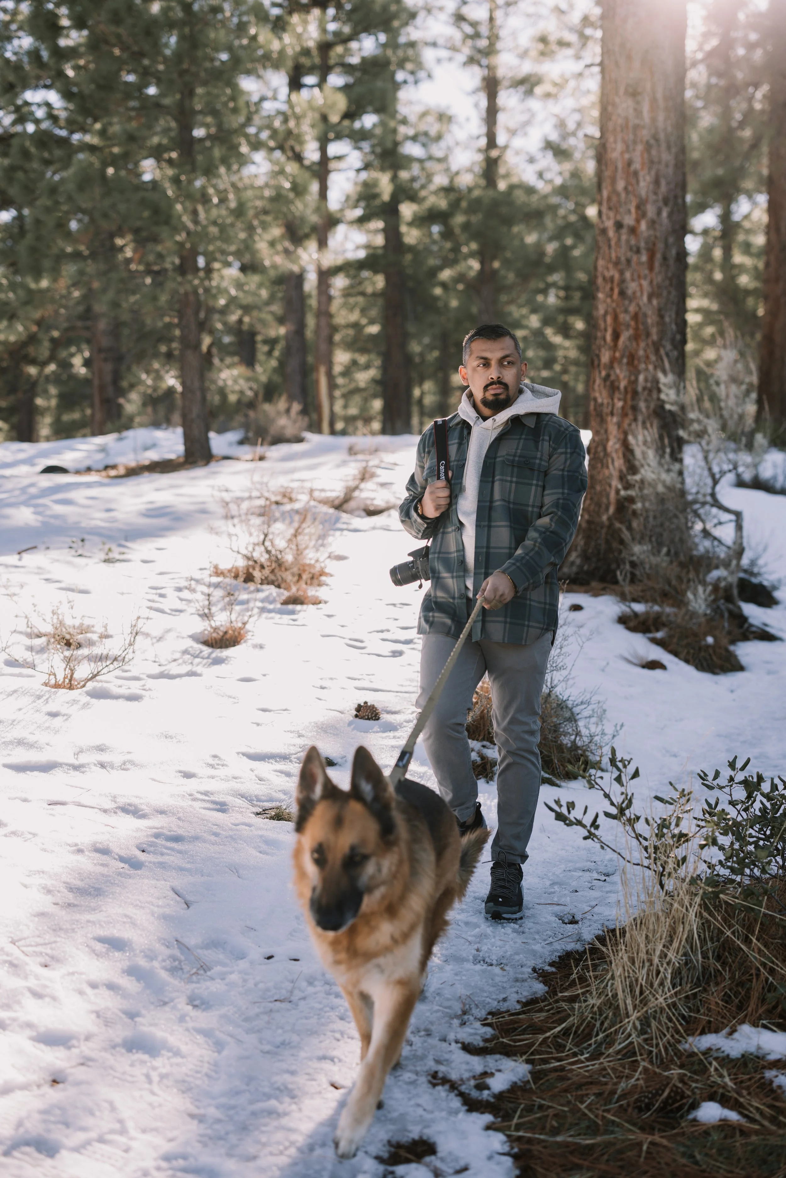 A man walking a German Shepherd dog in a snowy forest during daylight.