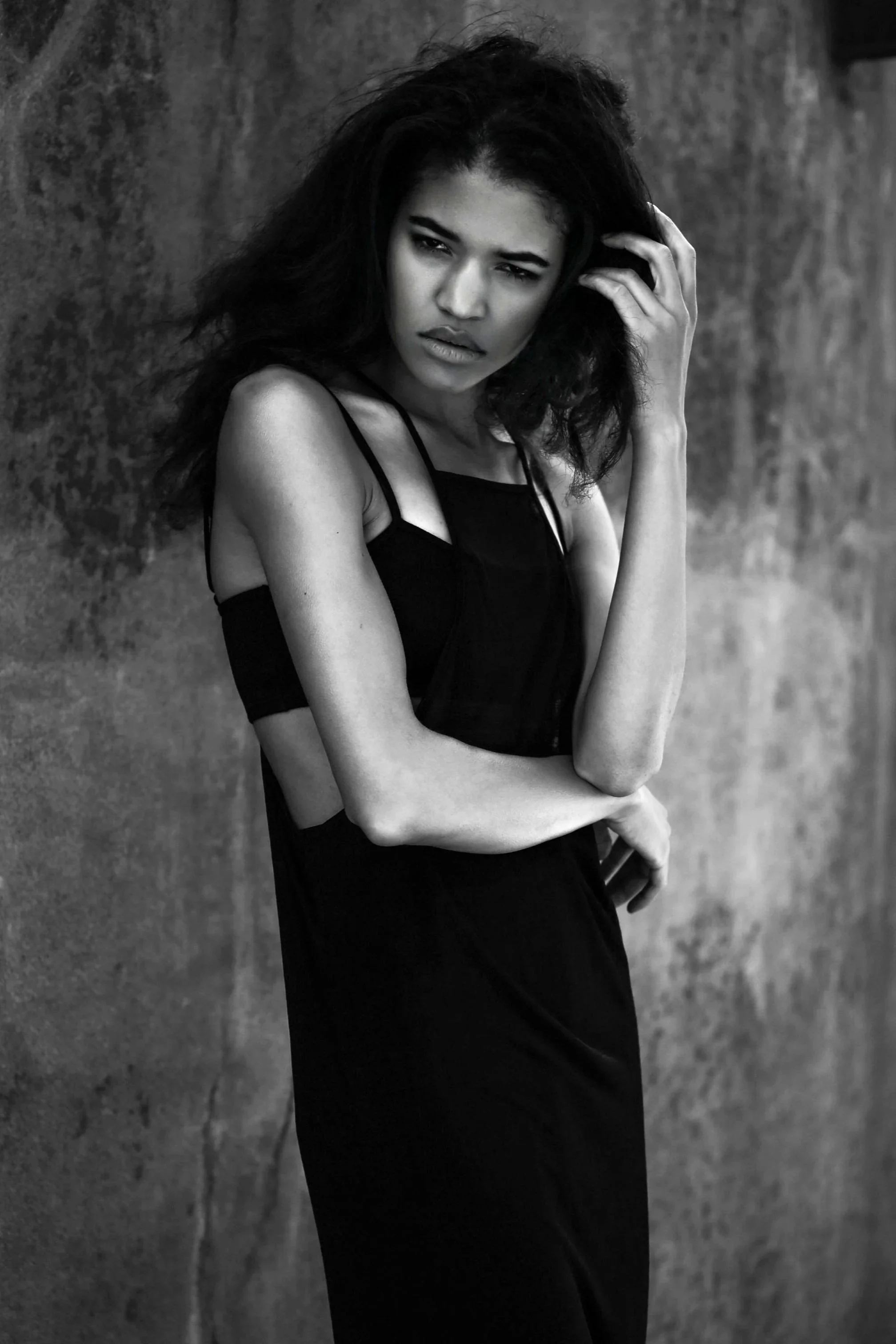 A black and white photo of a young woman with wavy hair, wearing a black dress with cutouts. She is standing against a textured wall, looking at the camera with a serious expression, with one hand in her hair and the other arm crossed.