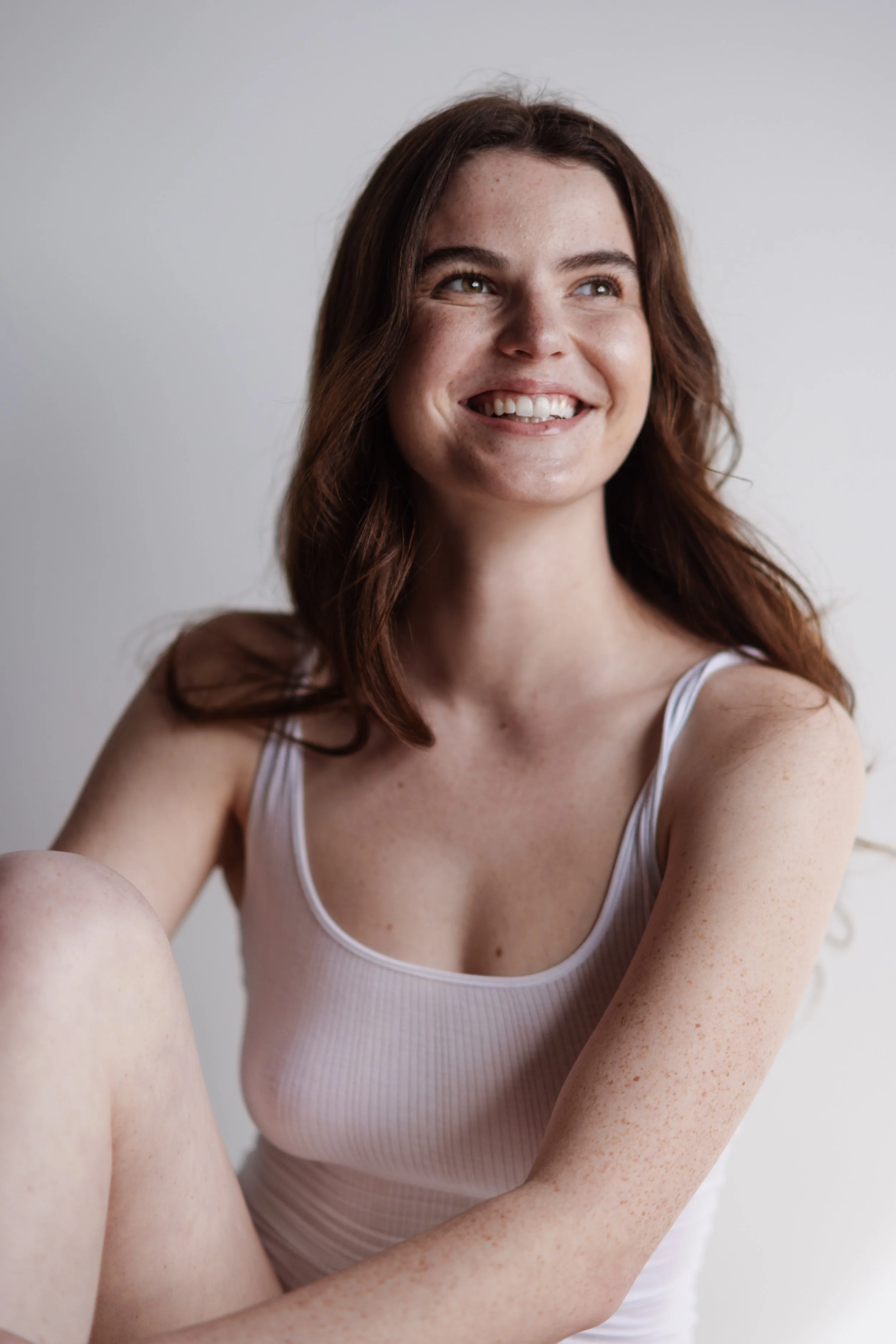 A young woman with wavy auburn hair, smiling, wearing a white tank top, sitting against a plain white background.