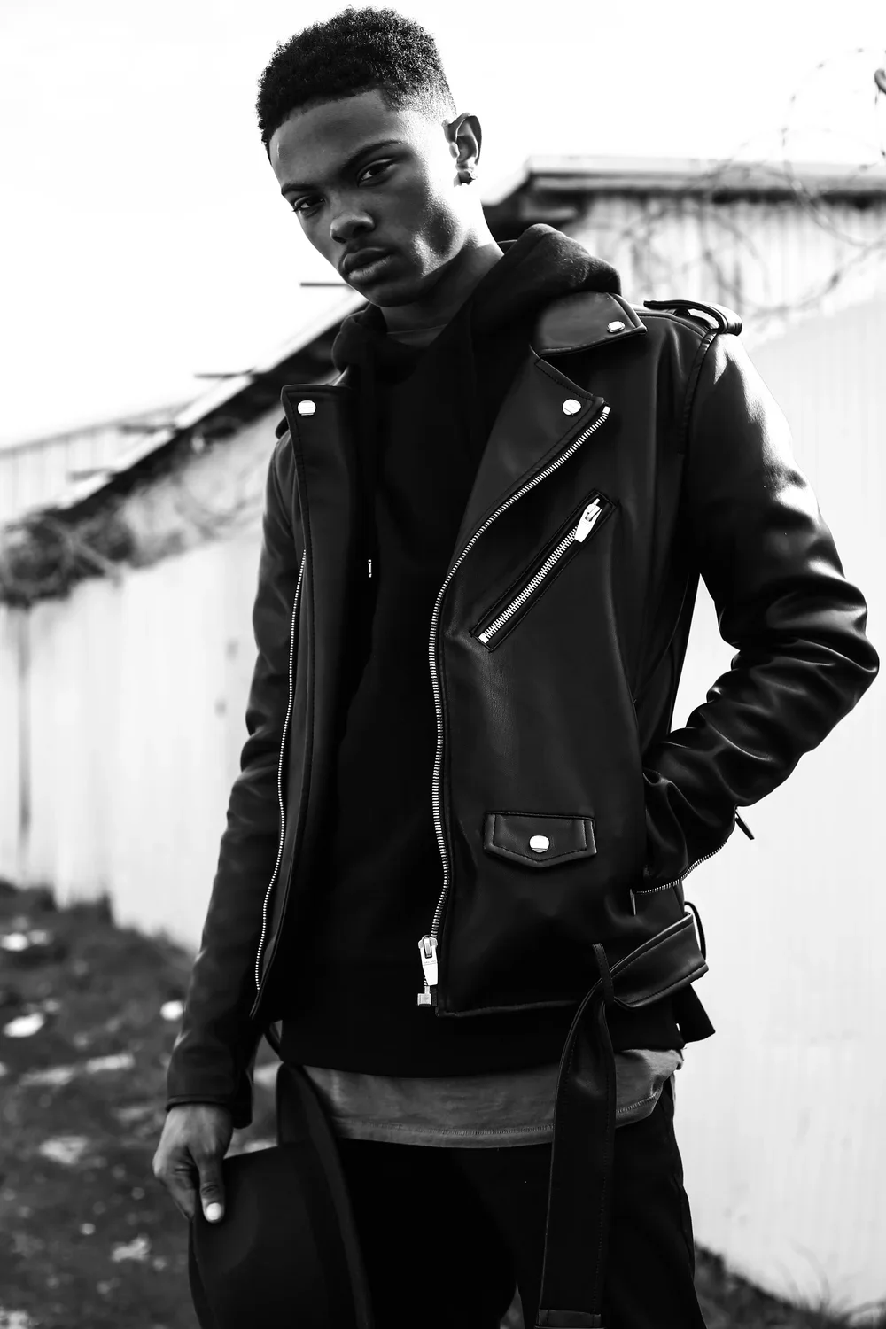 A young man with short curly hair wearing a black leather jacket with zipper details, a black hoodie underneath, and holding a hat, standing outdoors near a white fence, in black and white.