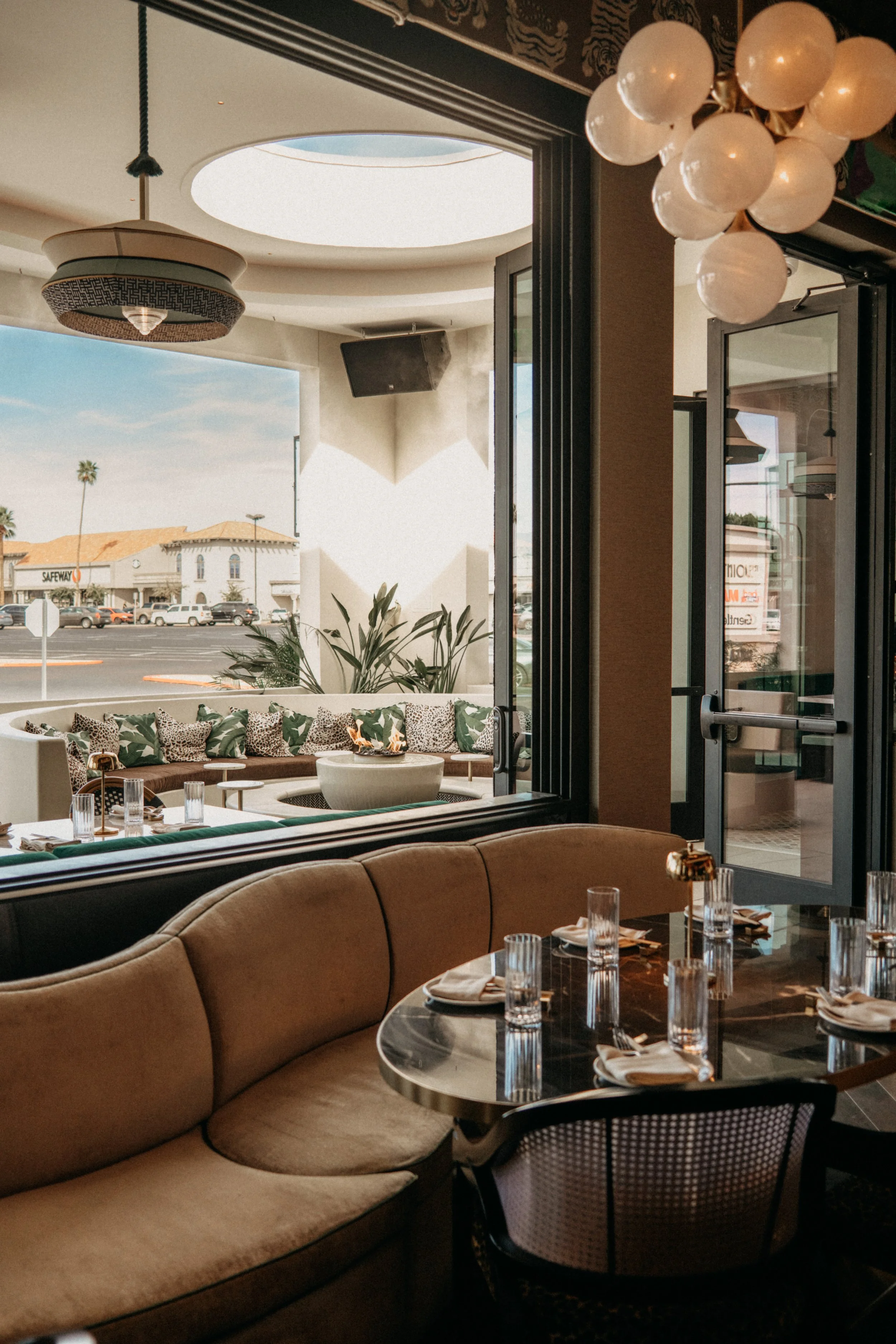 Interior of a restaurant or café with a beige banquette seating and a black marble table set with glasses, napkins, and utensils. Large windows reveal outdoor seating with pillows and greenery. Overhead, a modern chandelier and a cluster of white rou
