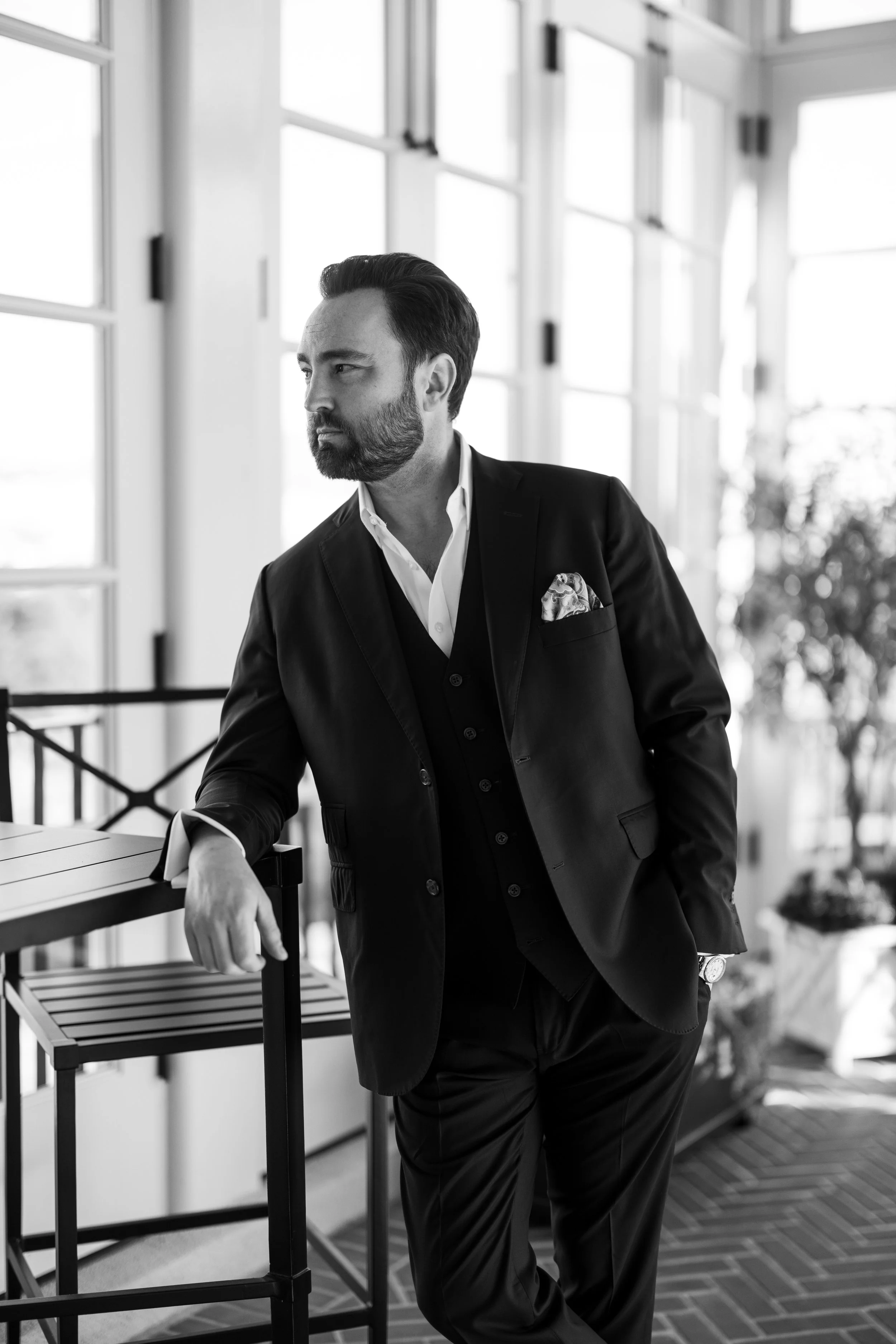 A well-dressed man with a beard and slicked-back hair leaning on a table in a bright, modern room with large windows.