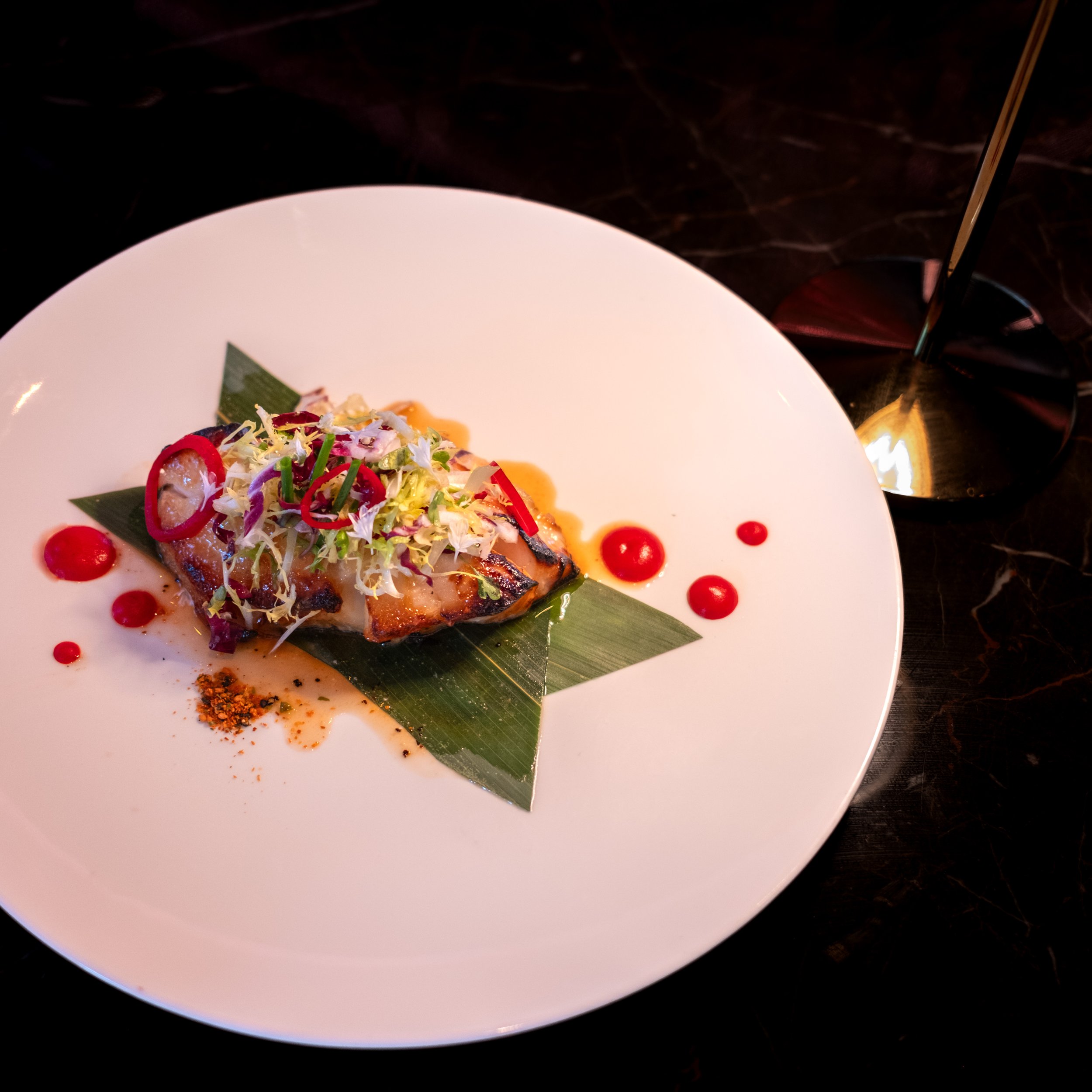 A plated dish with grilled fish topped with shredded vegetables and herbs, garnished with red chili slices, placed on a green leaf on a white plate with red sauce dots for presentation, next to a glass with a dark drink and a straw on a dark marble s