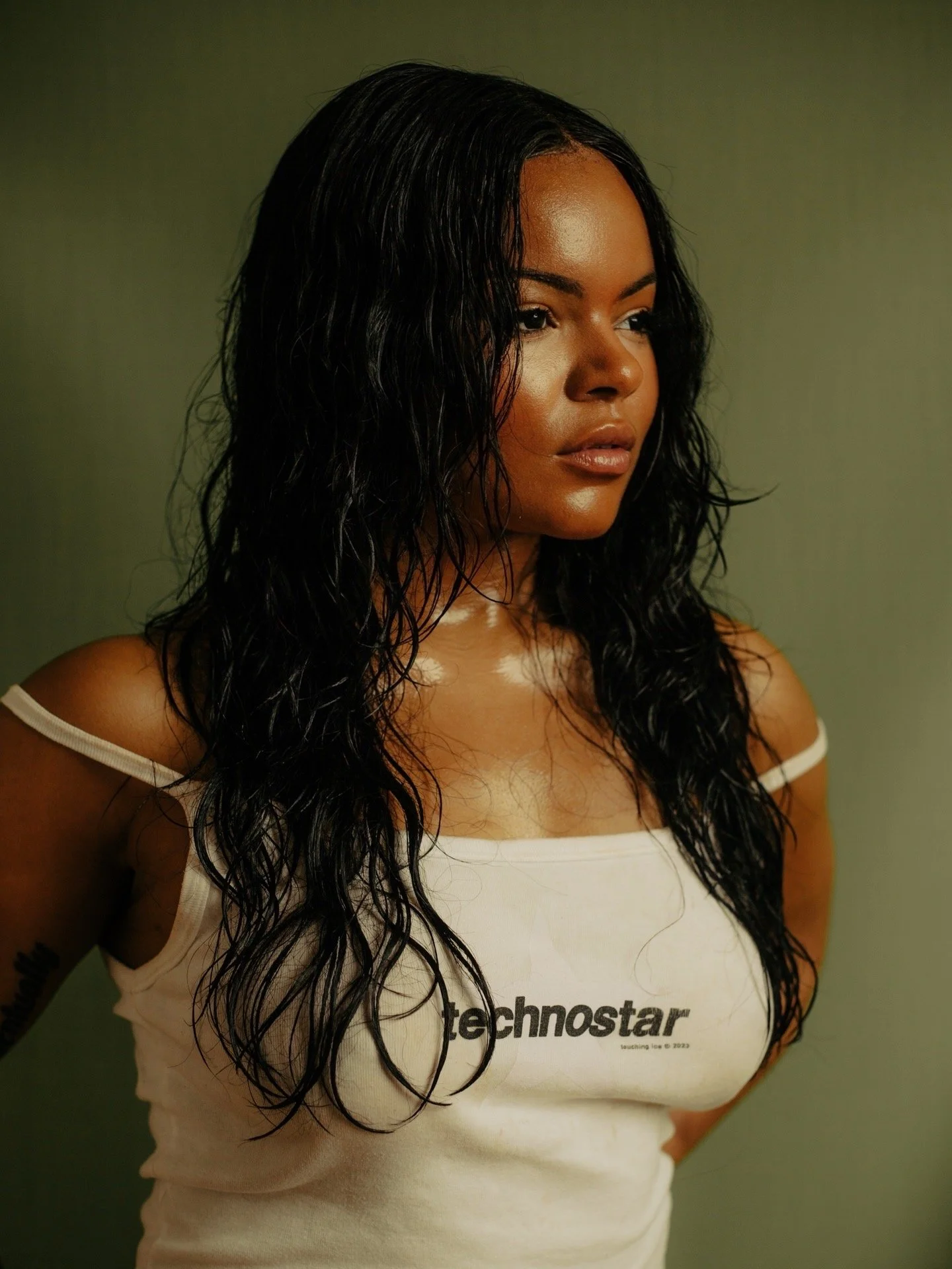 A young woman with long, wavy black hair, wearing a white tank top with the 'technostar' logo, looking to her right with a neutral expression against a green background.