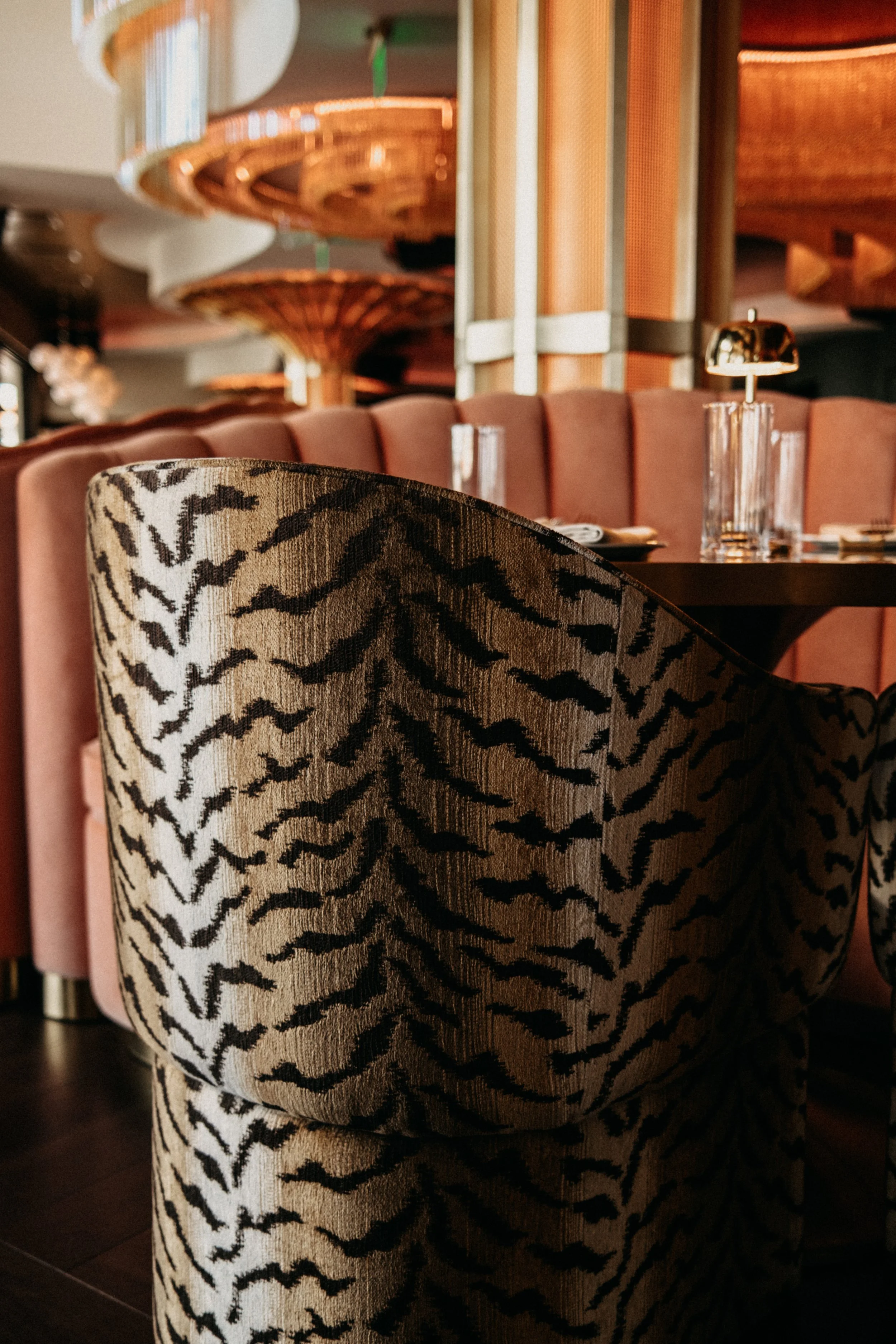 Close-up of a patterned upholstered chair in a restaurant with a table set with glassware and decorative lighting fixtures in the background.