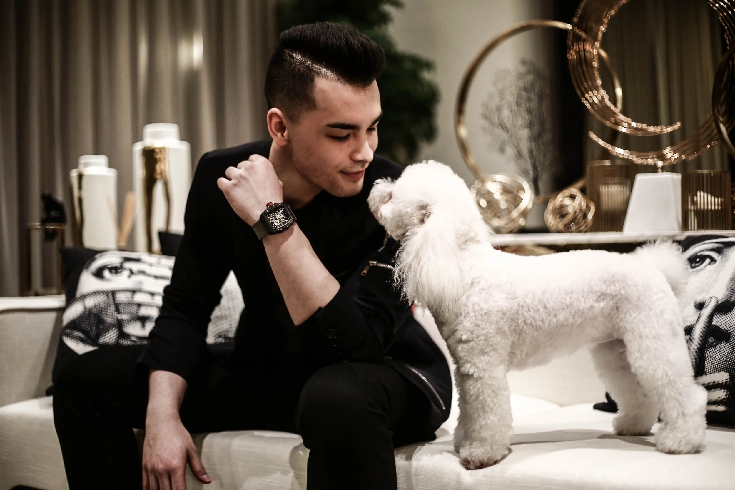 A young man with dark hair and a black watch sits on a white couch, leaning towards a white poodle. They are in a modern, stylish living room with gold decorative objects and framed artwork in the background.