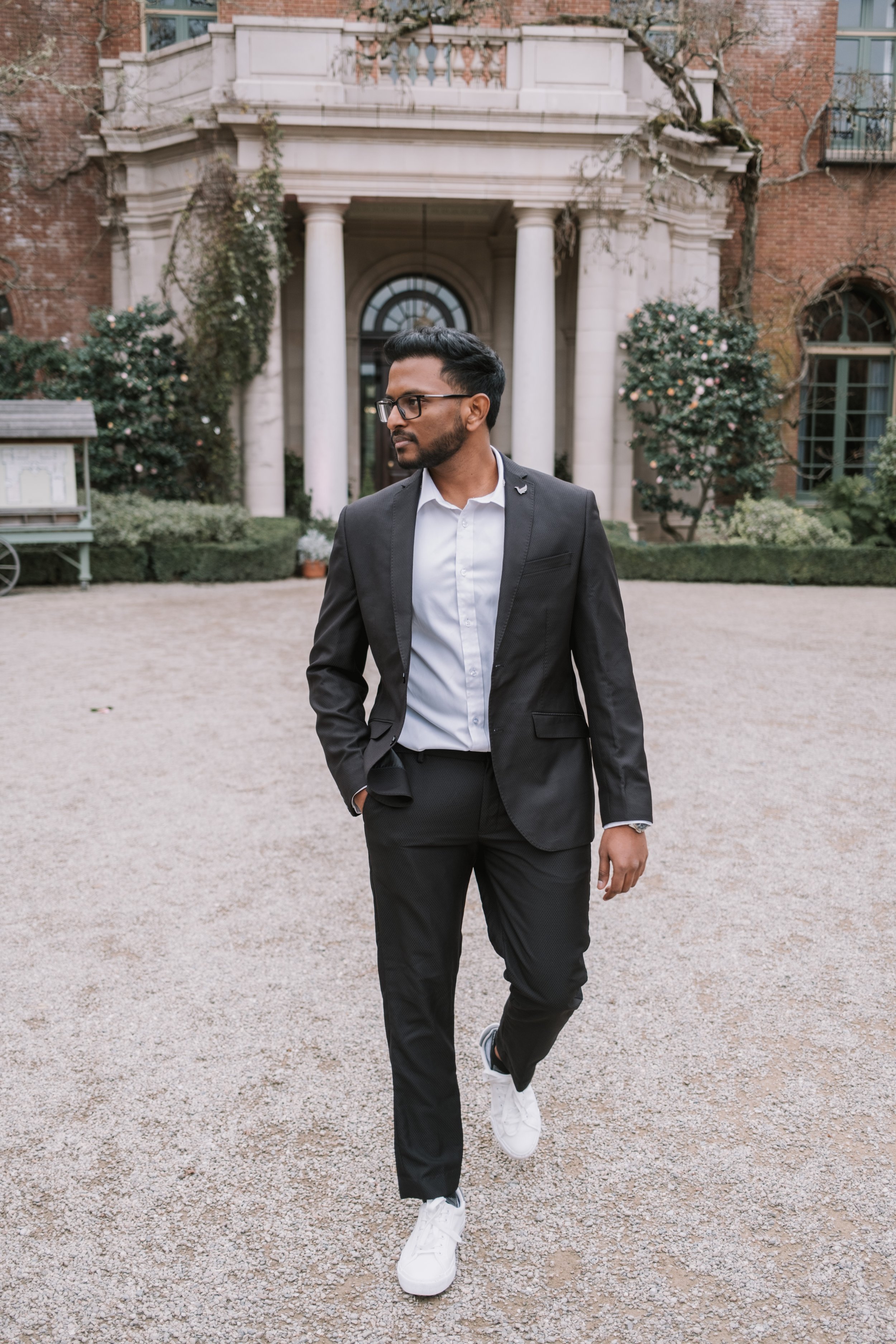A man in a black suit and glasses walking on a gravel path in front of a historic building with columns and trees.