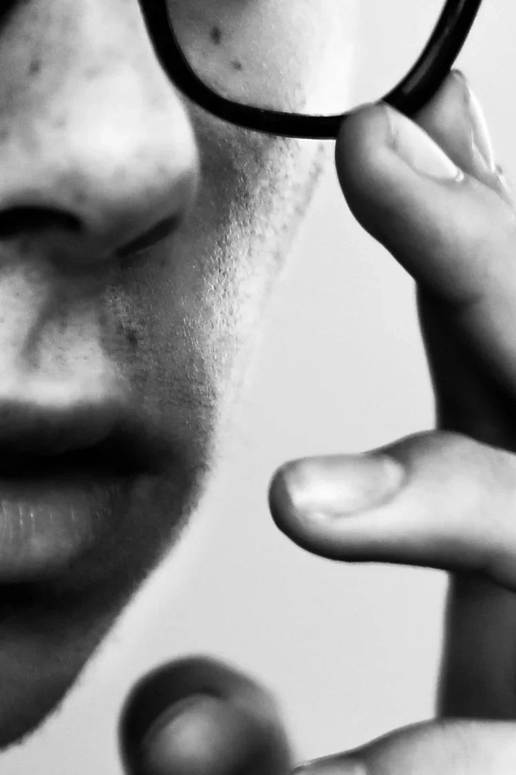 Close-up of a person's face with lips and nose visible, holding a small mirror near the eye, shown in black and white.