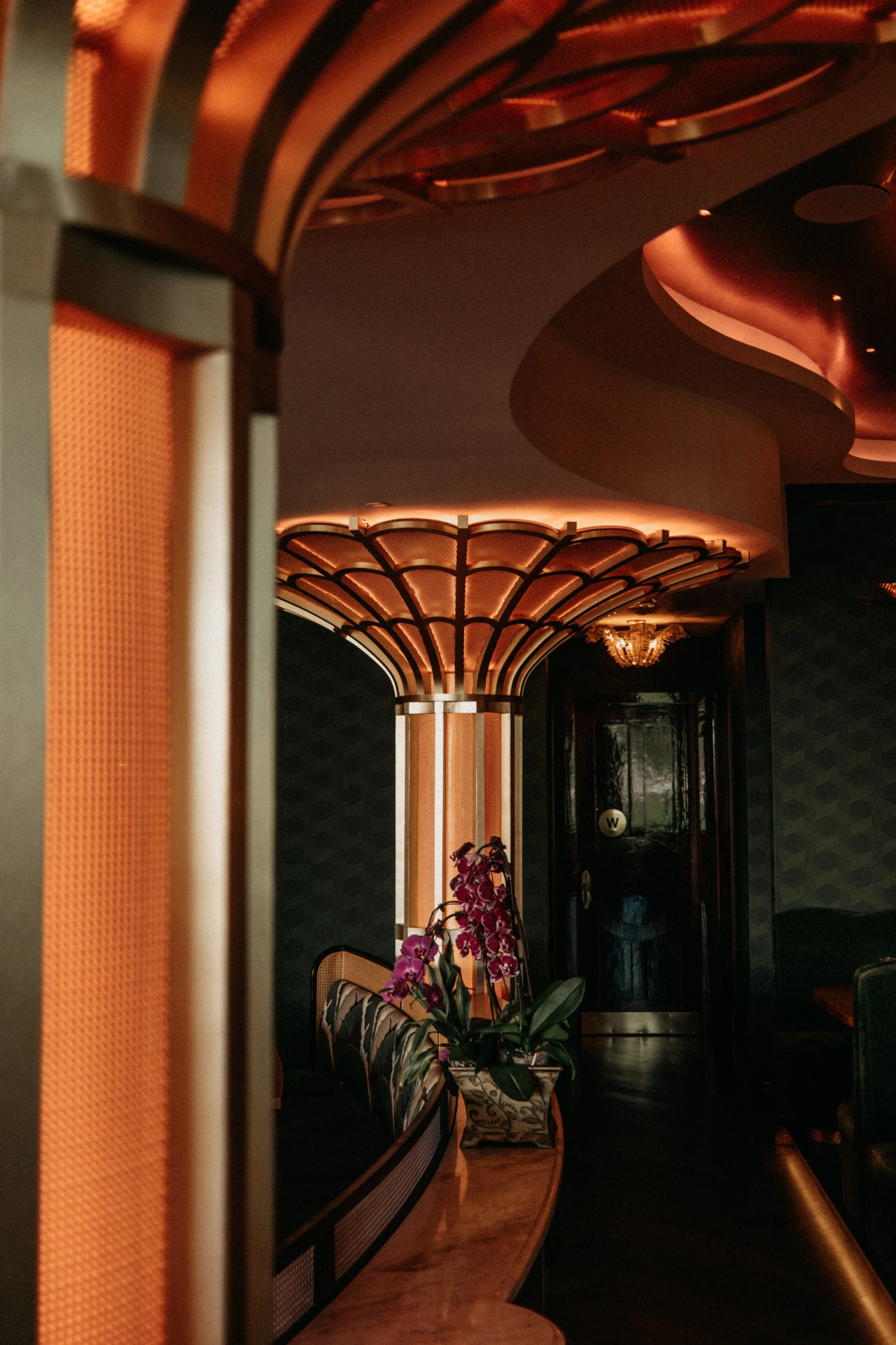 Interior of a luxurious hotel lobby with ornate lighting fixtures, a floral arrangement of purple orchids, dark walls with patterned wallpaper, and elegant seating areas.