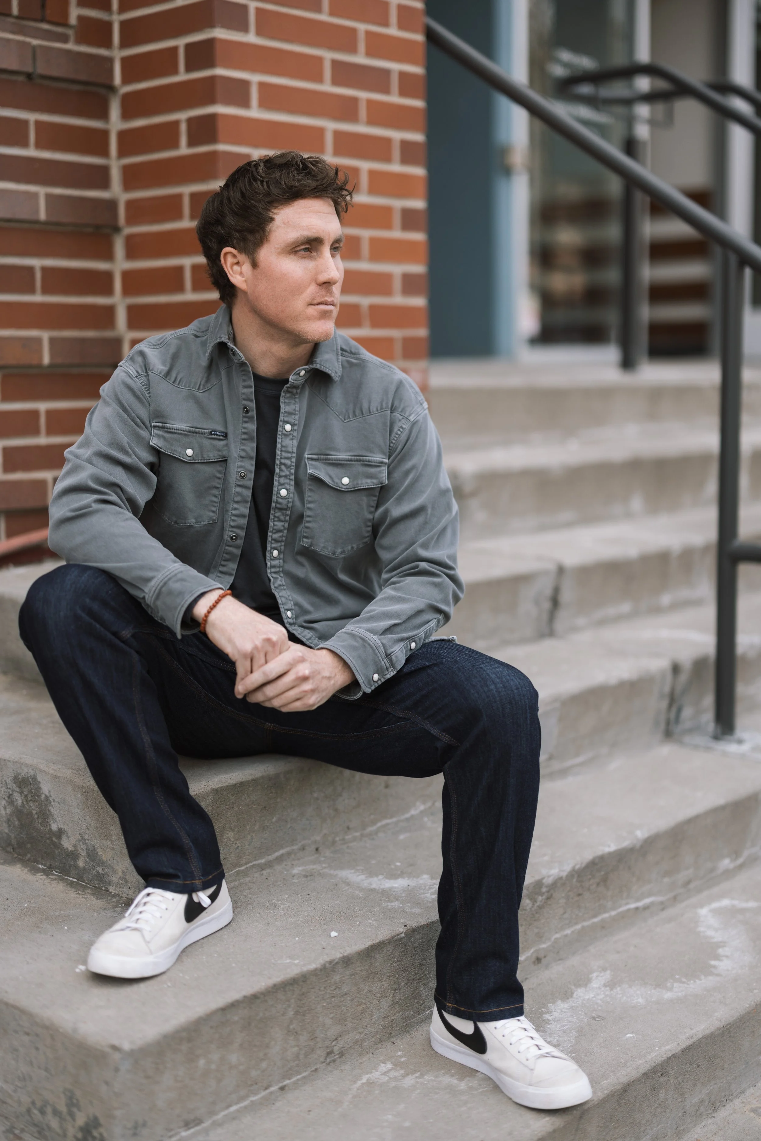 A young man sitting on concrete steps outside a brick building, looking to his left with a serious expression, wearing a gray jacket, black jeans, and white sneakers.