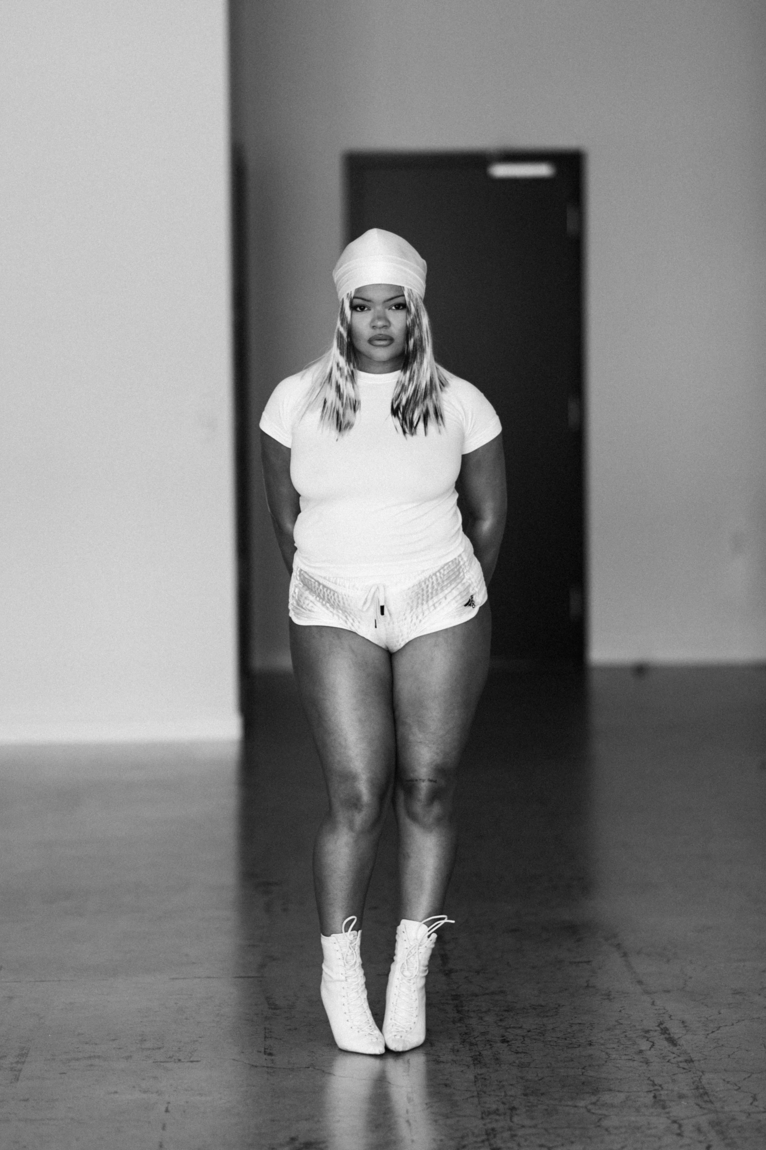 A woman with long hair, wearing a head wrap, white t-shirt, shorts, and high-heeled shoes, standing alone in an indoor space, looking directly at the camera.