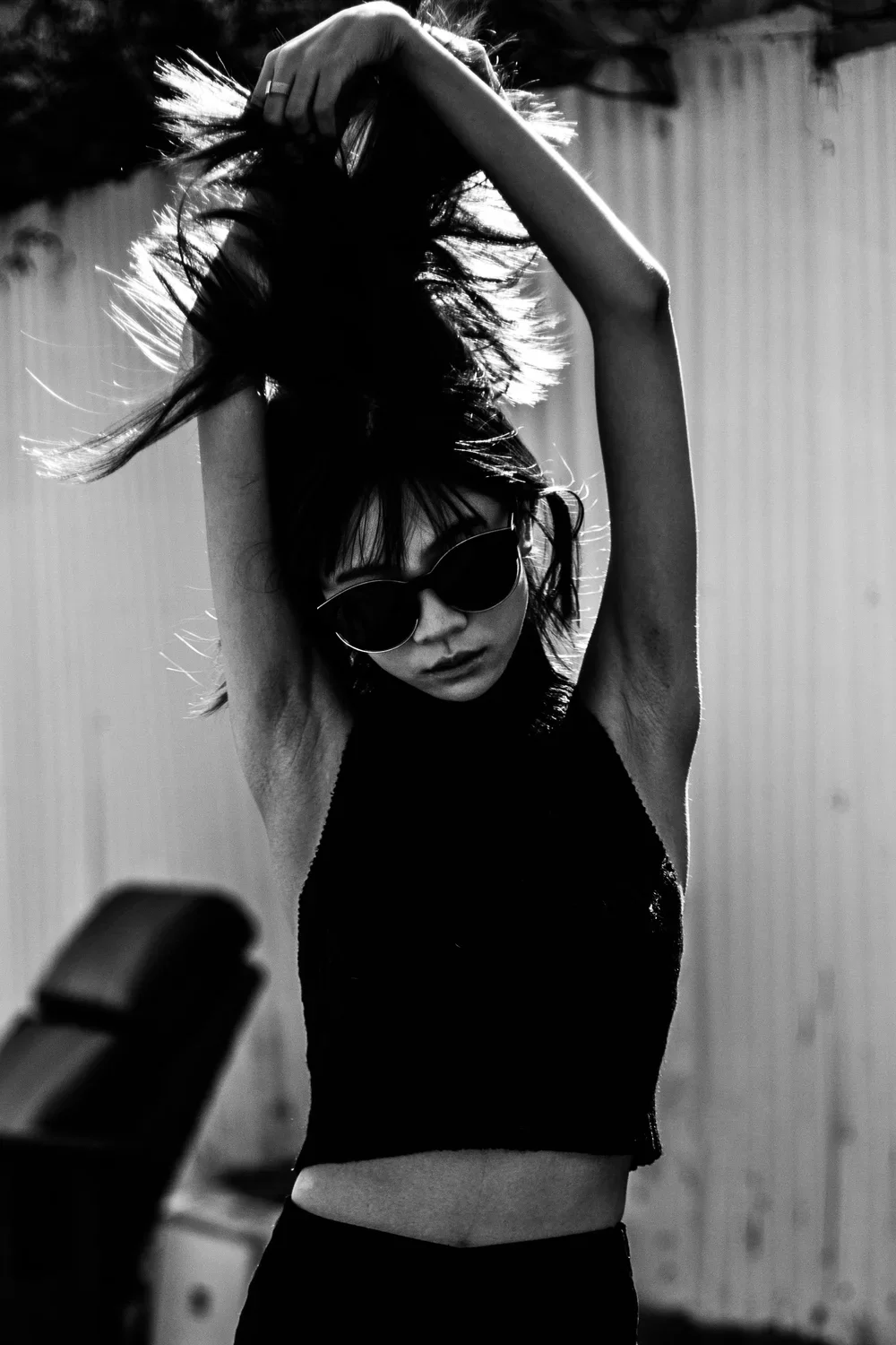 A woman with dark hair, sunglasses, and a sleeveless top is touching her hair with both hands, standing in an outdoor setting with a background of a wooden fence.