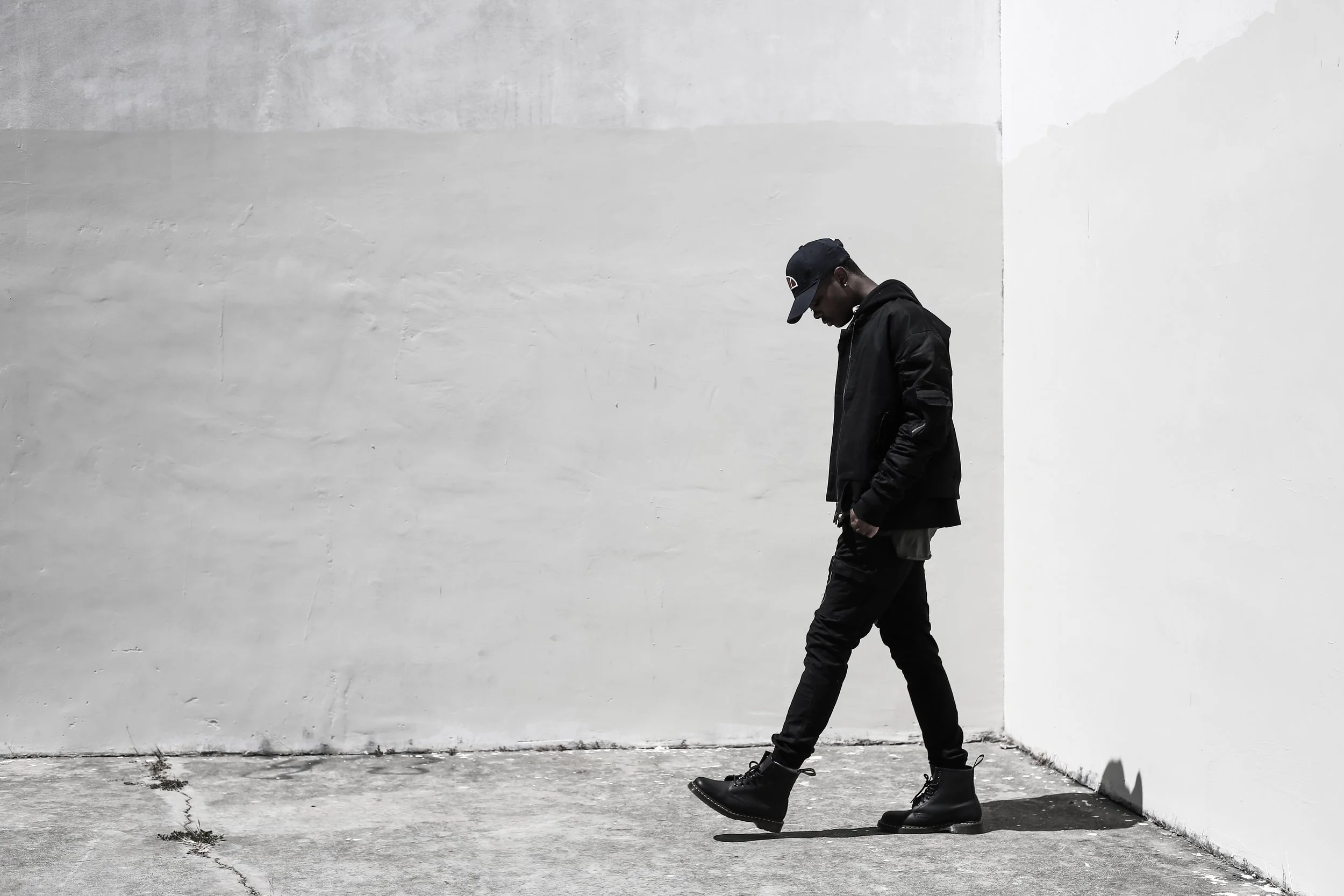 Young man in black jacket, pants, and boots walking on concrete sidewalk beside a white wall, wearing a black cap, with head bowed and hands in pockets.