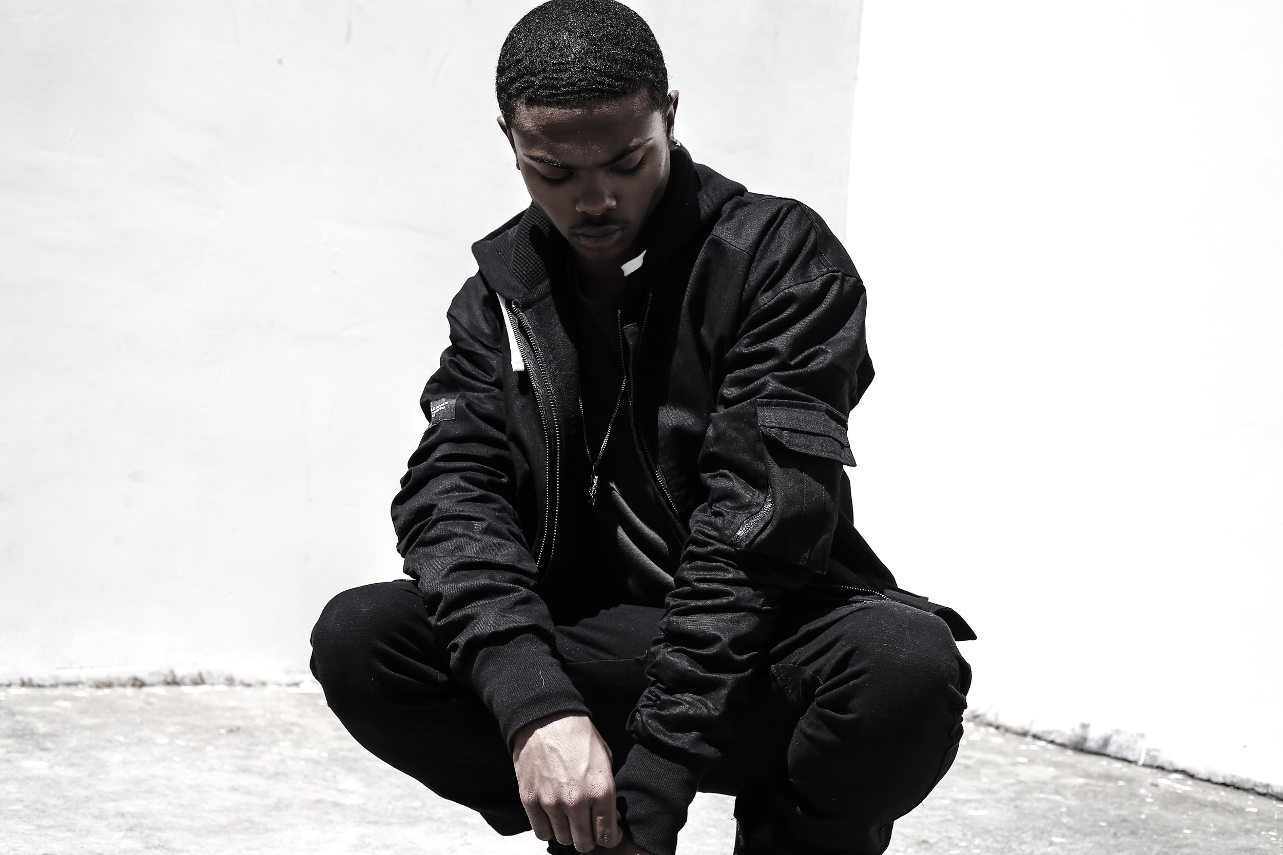 Young man in black jacket and black pants squatting against a plain white wall, looking down.
