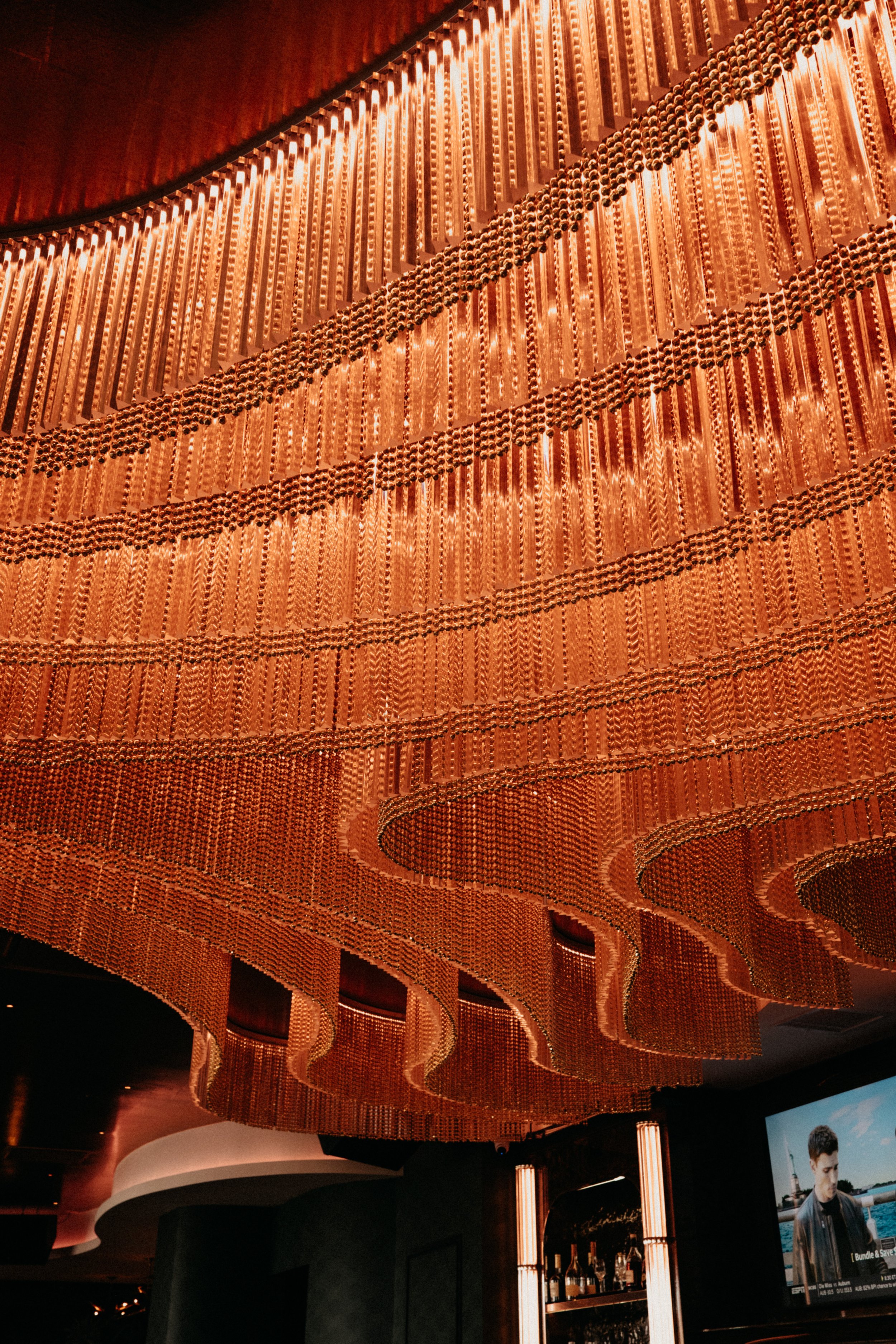A large, elaborate chandelier made of copper-colored tubes and beads hanging from a dark ceiling in an upscale indoor setting.