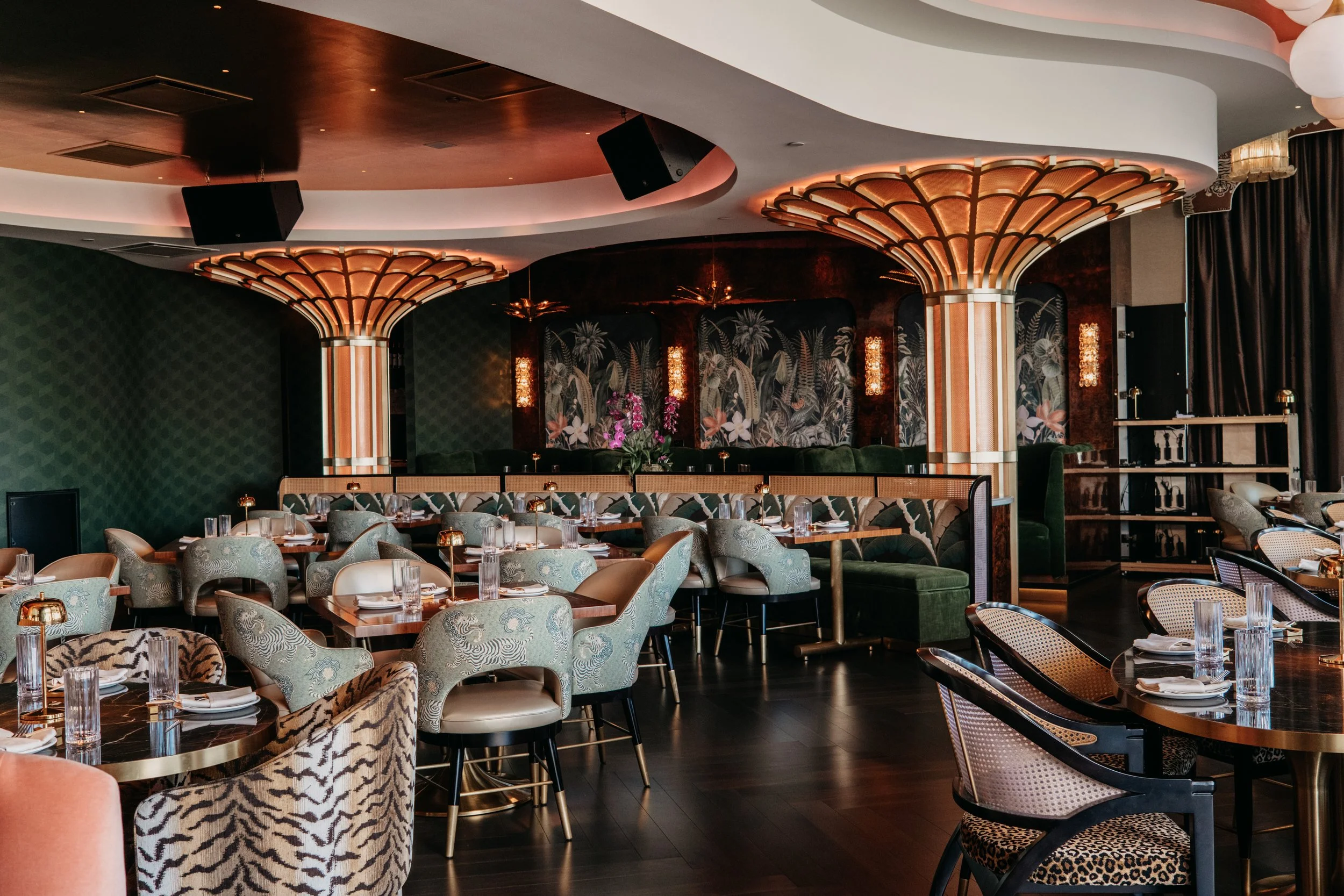 Elegant restaurant interior with patterned chairs, decorative lighting, dark wood floors, and lush wallpaper featuring botanical designs.